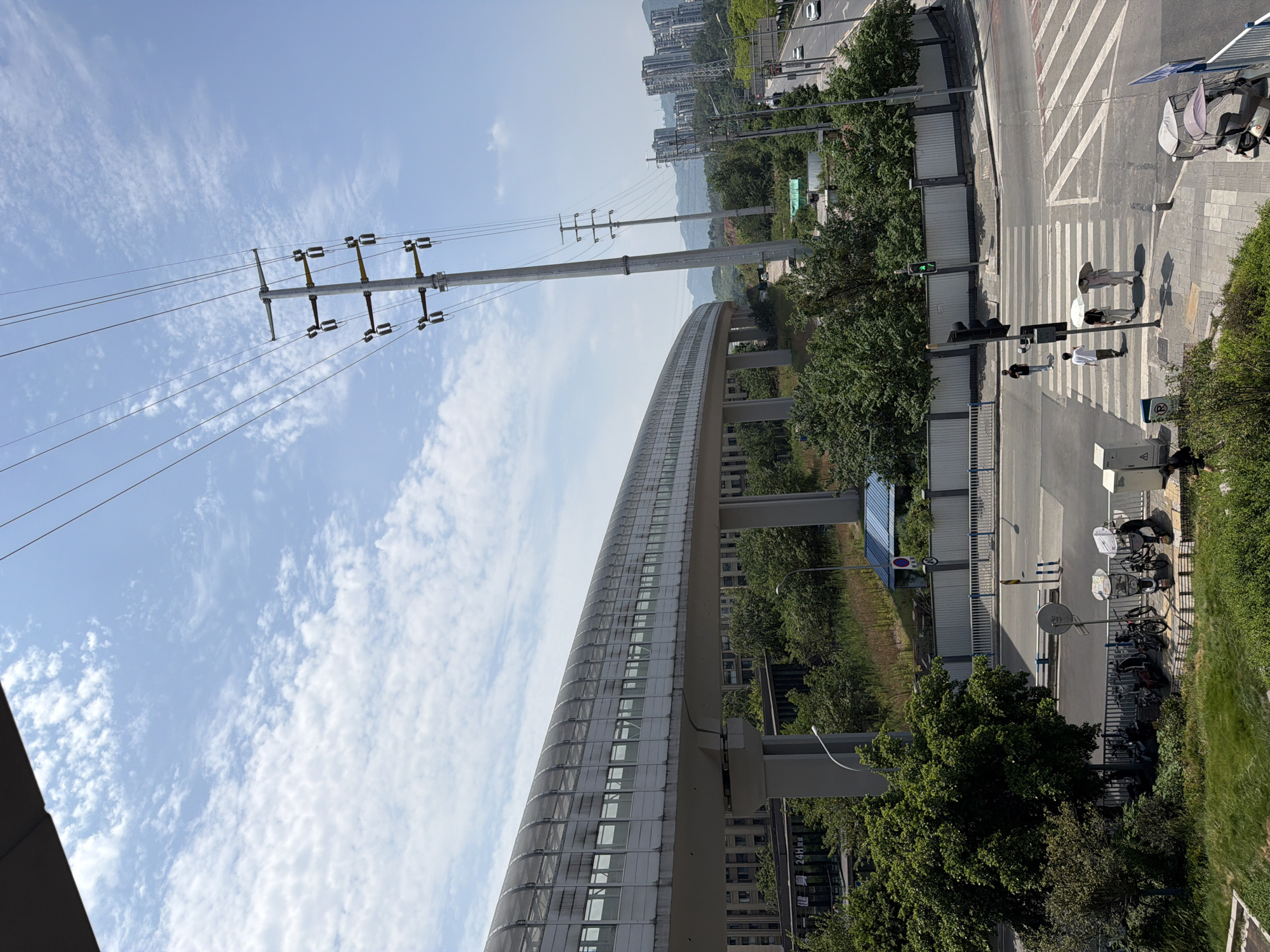 The image captures a street scene in Tiaodeng, China, on a sunny day with scattered clouds. Dominating the left side of the frame is a section of an elevated, enclosed walkway or monorail track, supported by substantial pillars. This structure curves away from the viewer and appears to be made of white panels and a transparent, mesh-like roof. Adjacent to the walkway are numerous trees and some low-rise buildings.
To the right of the elevated structure, a tall, grey power line pylon stands prominently, carrying multiple high-voltage wires. Further in the distance, a cluster of modern, high-rise apartment buildings is visible against a hazy mountain backdrop.
In the foreground, a busy intersection with a crosswalk is depicted. Several people are crossing the street. Some are walking with umbrellas, suggesting protection from the sun. Parked bicycles and electric scooters are visible along the sidewalk. Traffic lights indicate the flow of vehicles, and a few cars are seen on the road. The overall impression is of an urban environment integrating modern infrastructure with elements of nature.
