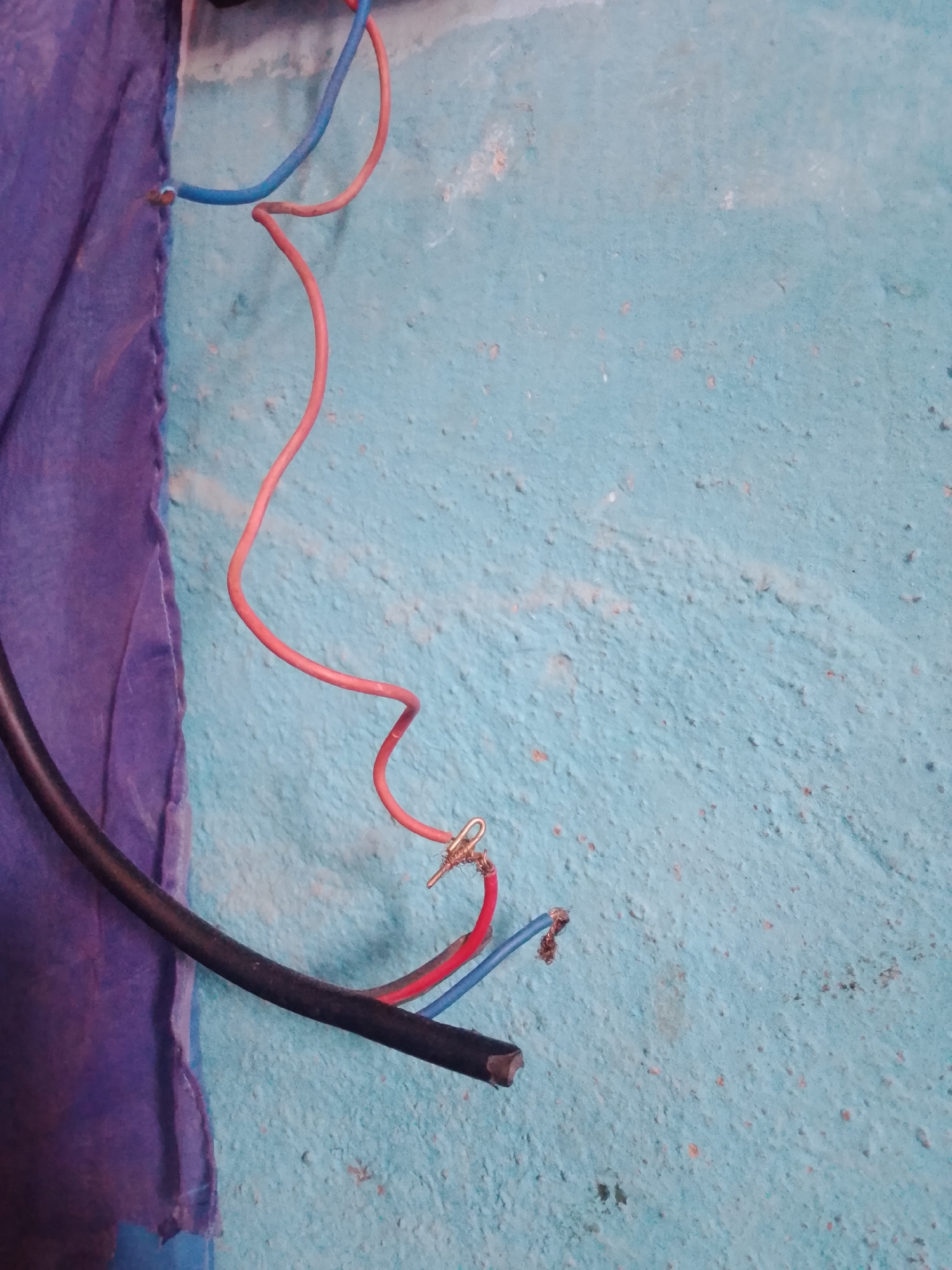 The media file displays a close-up shot of electrical wires against a textured, blue wall. On the left side of the image, a portion of a purple cloth, possibly fabric, is visible, with a rough edge and some frayed threads. Several electrical wires are present, including a thick black cable, a red wire, and a blue wire. The red and blue wires appear to be stripped at the ends and are connected to something with a small, metallic fastener. One of the red wire's ends is twisted around this fastener, and the blue wire's end is also partially visible. The wires are arranged in a somewhat haphazard manner, suggesting a work in progress or a disarray of electrical connections. The blue wall has a rough, uneven surface with some darker speckles and lighter patches, hinting at its texture and possibly age. No people or discernible text are present in the image. The lighting suggests an indoor environment, with no specific indicators of time of day or weather conditions. The focus is primarily on the electrical components and the wall surface.