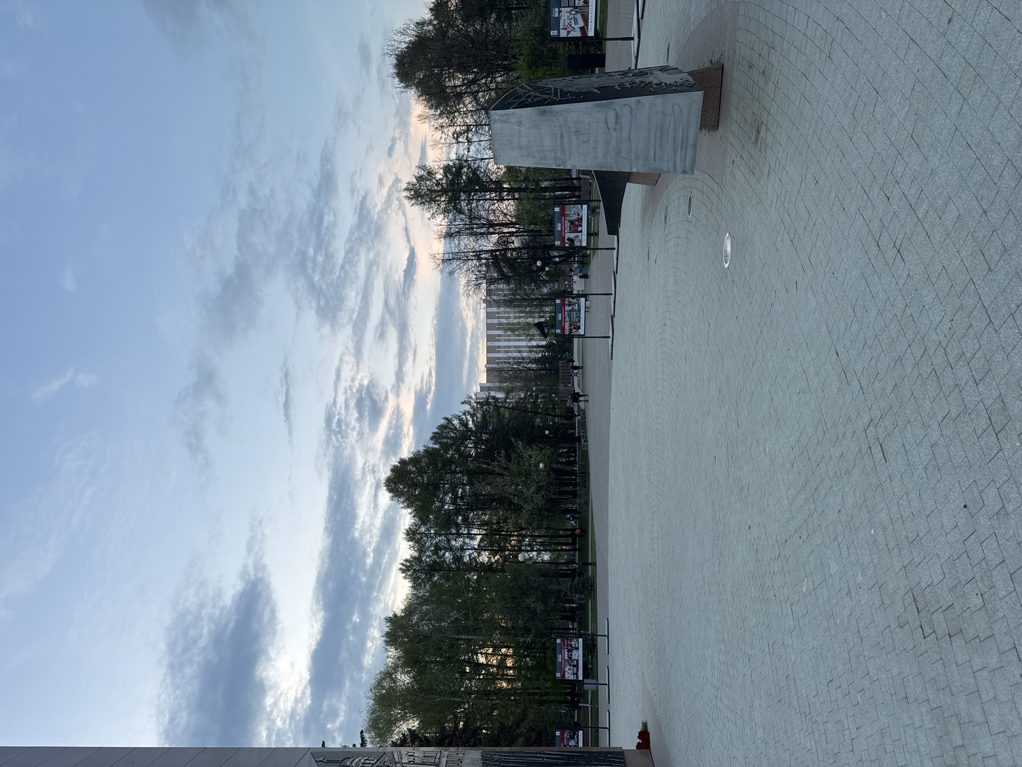 The image shows a public space in Izhevsk, Russia, likely a memorial or monument area. The sky occupies the upper half of the image with scattered clouds. A line of trees divides the sky from the paved ground area. Along the tree line are lampposts and what appear to be informational signs or display panels. A concrete or stone monument stands to the left of the tree line. The ground is paved with small grey stones.