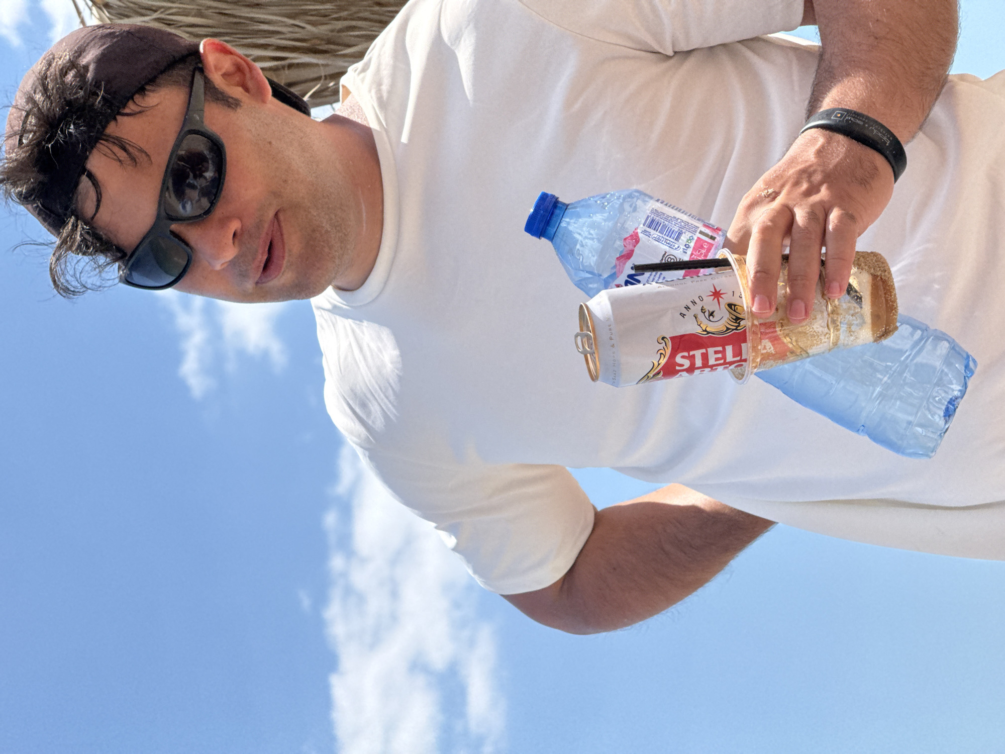 A man wearing a white t-shirt, dark sunglasses, and a baseball cap holds a plastic cup with a straw and a can of Stella Artois. A partially filled plastic water bottle with a blue cap is also in his hand. He stands against a clear blue sky with sparse clouds. The background features a hint of thatched material, suggesting an outdoor setting, possibly a beach umbrella or a thatched roof. The man's left hand rests on his hip, and he looks off to the side with a relaxed expression. The scene is illuminated by bright sunlight, casting subtle shadows. The presence of the drinks and casual attire suggests a leisure or vacation setting. The image does not contain enough information to definitively place the scene in Kranevo, Bulgaria.