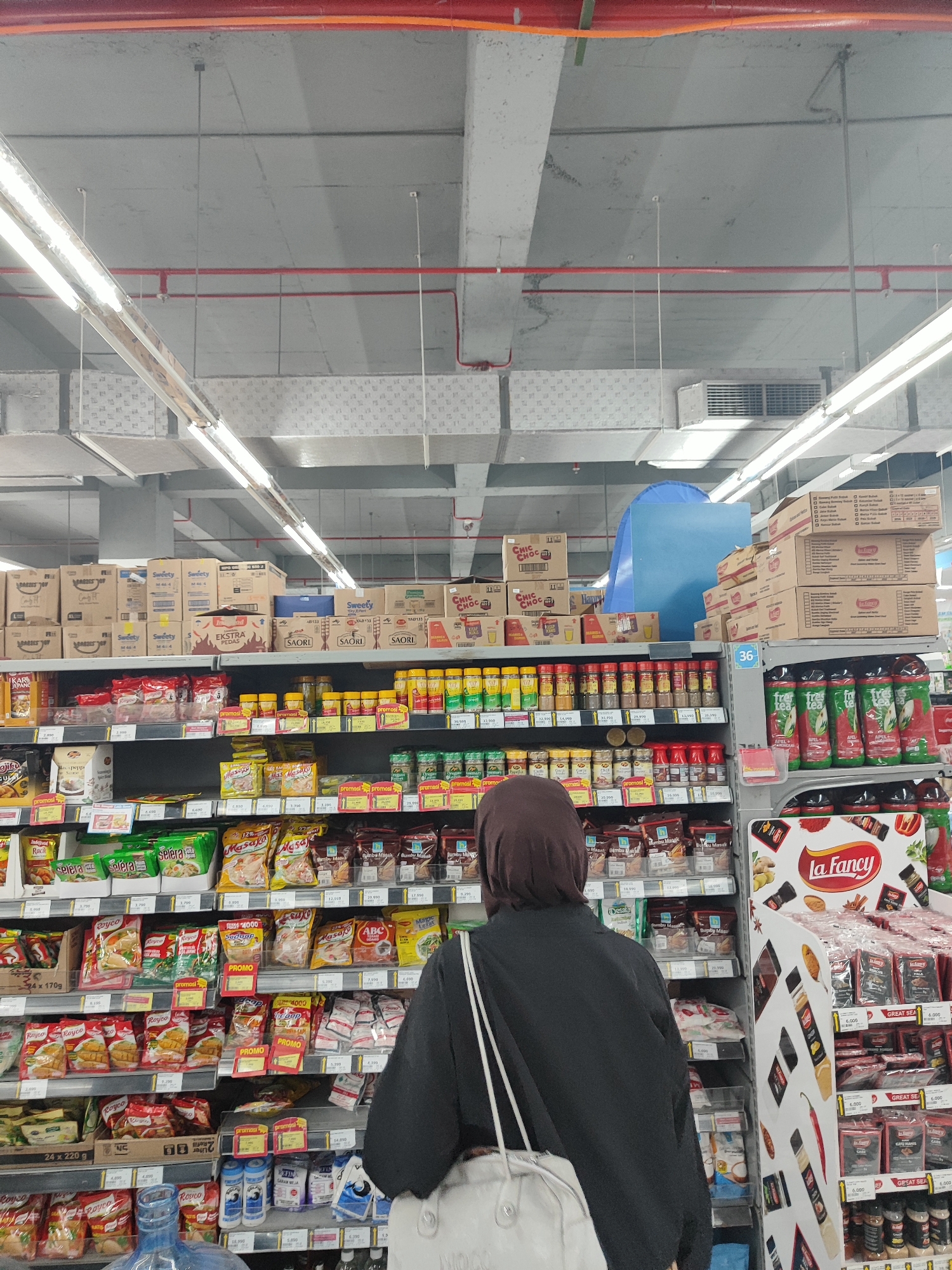 A rear view of a person wearing a hijab and a dark jacket is visible in the foreground, facing away from the camera. They are carrying a white bag and are in a supermarket aisle. The shelves are stocked with various food items, including packaged goods, spices, and drinks. The aisle has fluorescent lighting overhead, and the ceiling is made of concrete with exposed pipes and ventilation systems. Boxes are stacked on higher shelves, and some shelves have price tags or promotional labels. A blue tarp-like object is visible on a higher shelf in the background. The scene appears to be in a commercial establishment, likely a grocery store, within the city of Nangkaan, Indonesia.
