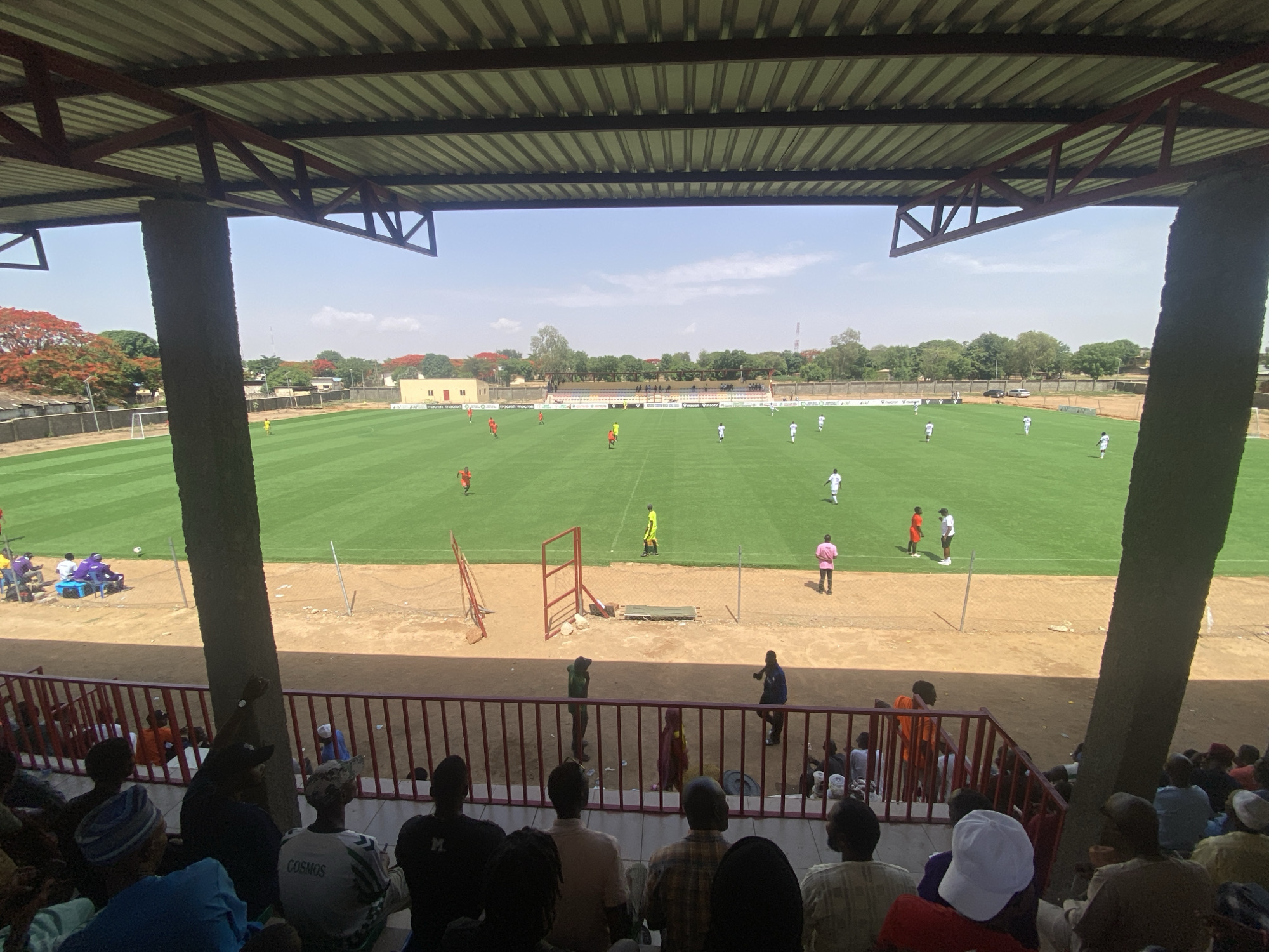 A soccer match is in progress in Tudun Wada, Nigeria. The game is taking place on a green field, observed by fans from a stadium's covered seating area. Players in white and orange uniforms are actively participating in the match. A soccer goal is visible in the background on the left. Between the spectators and field is open ground and a fence. The stadium's support beams are visible on either side.