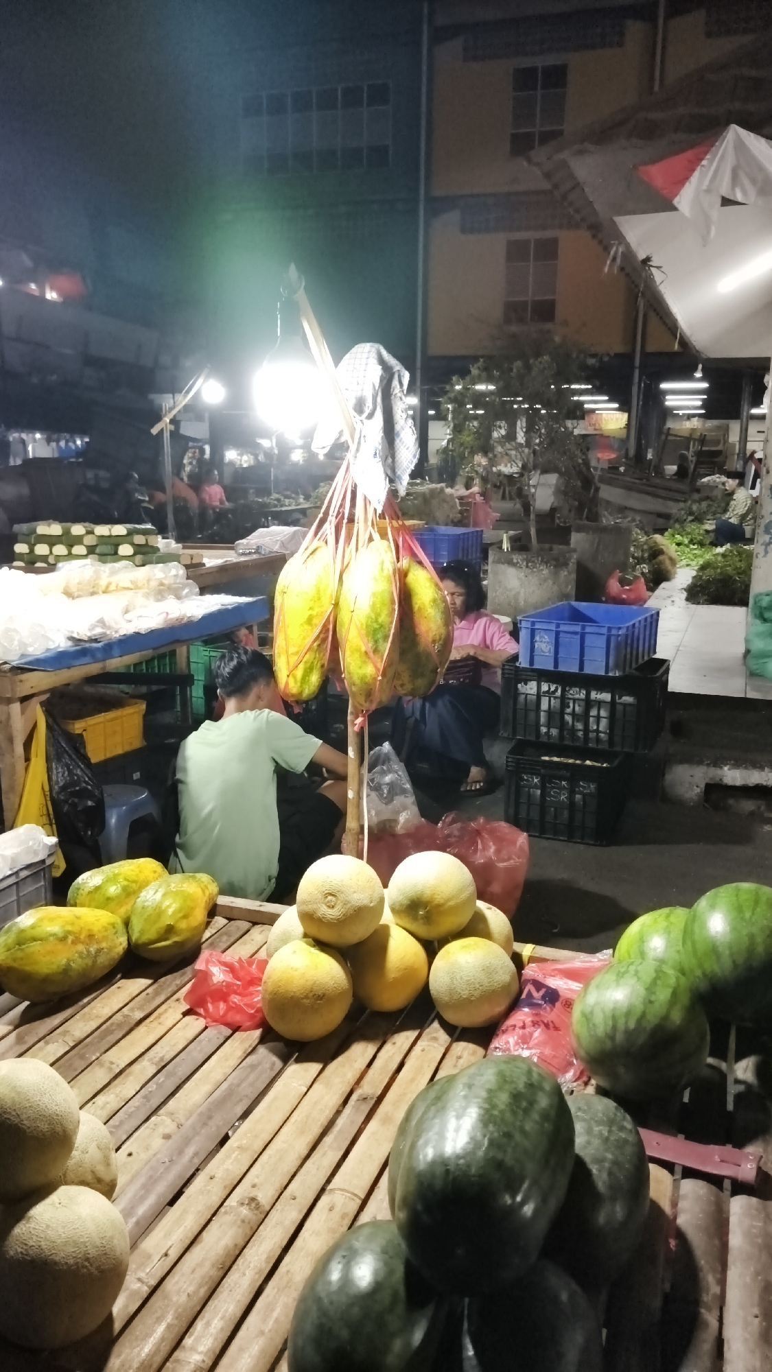 This is a nighttime outdoor market scene in South Jakarta, Indonesia, likely in a commercial district or near residential areas where informal markets operate after dark. The market is illuminated by artificial lighting, including a bright overhead lamp that casts a strong glow. In the foreground, there is a display of fresh produce on a wooden stall. Several watermelons are stacked, some with their rinds visible. Next to them, piles of melons, likely cantaloupe or honeydew, are arranged. Further back on the stall, a row of papayas is laid out, and a bunch of papayas are hanging from a makeshift wooden support, tied with red string.
In the middle ground, two individuals are visible. A young man wearing a light green t-shirt is seated behind the papaya display, appearing to be working or attending to the produce. To his right, a woman wearing a pink top and dark pants is seated, possibly a vendor, with her hands near a bag. Behind them, various market structures and goods are present. Stacks of blue and black plastic crates are visible, suggesting produce transportation or storage. In the background, buildings with windows are dimly lit, indicating commercial or residential structures. The overall environment suggests a bustling, albeit slightly chaotic, nighttime marketplace common in urban areas of Indonesia.