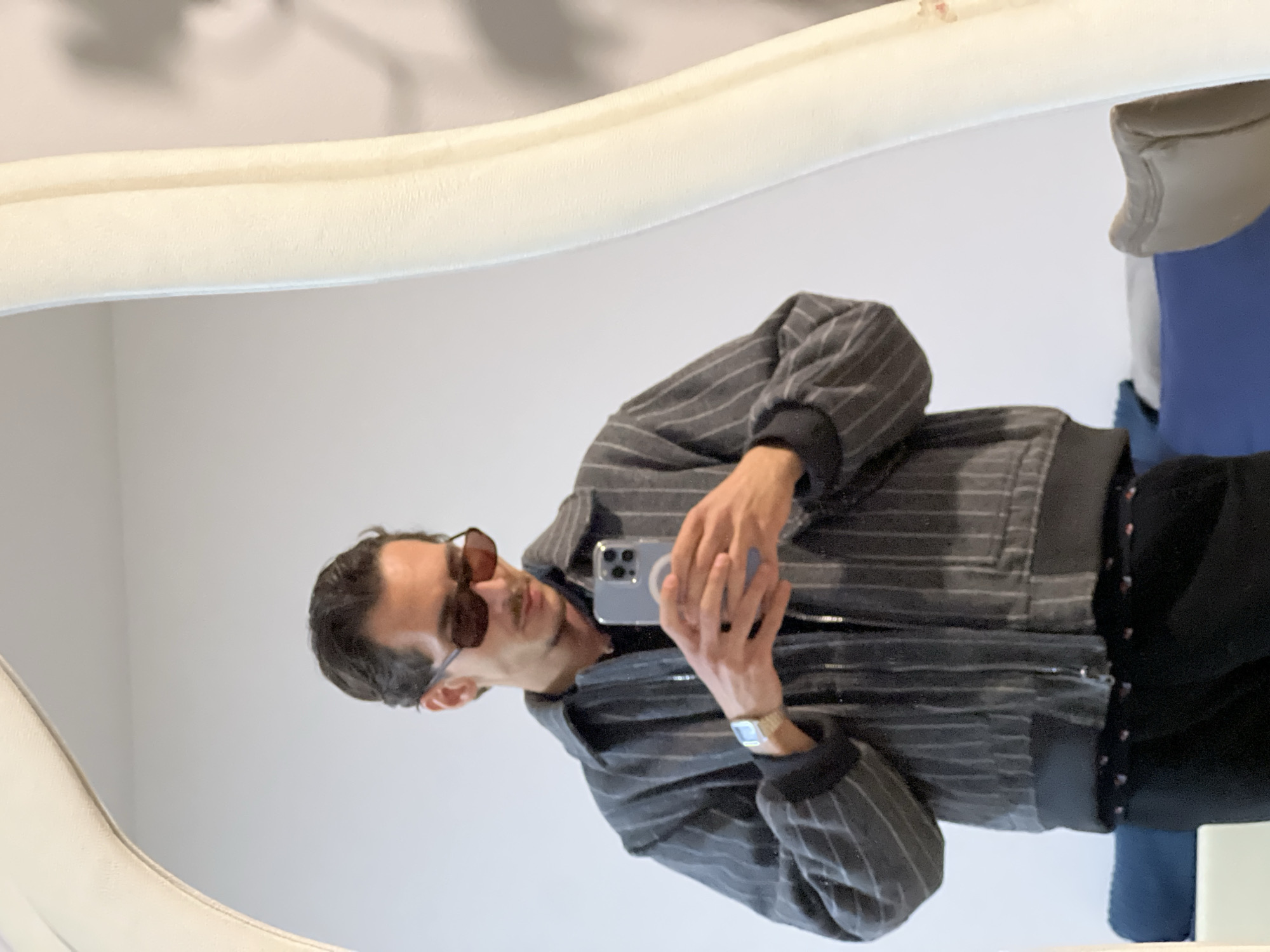 A man is reflected in a mirror. He is wearing a gray striped jacket, sunglasses, and a watch. He is holding a smartphone in his hands, presumably taking a picture of himself in the mirror. The mirror has a white frame. The background is a plain white wall. The location is not discernible as Boreti, Montenegro, without further information.