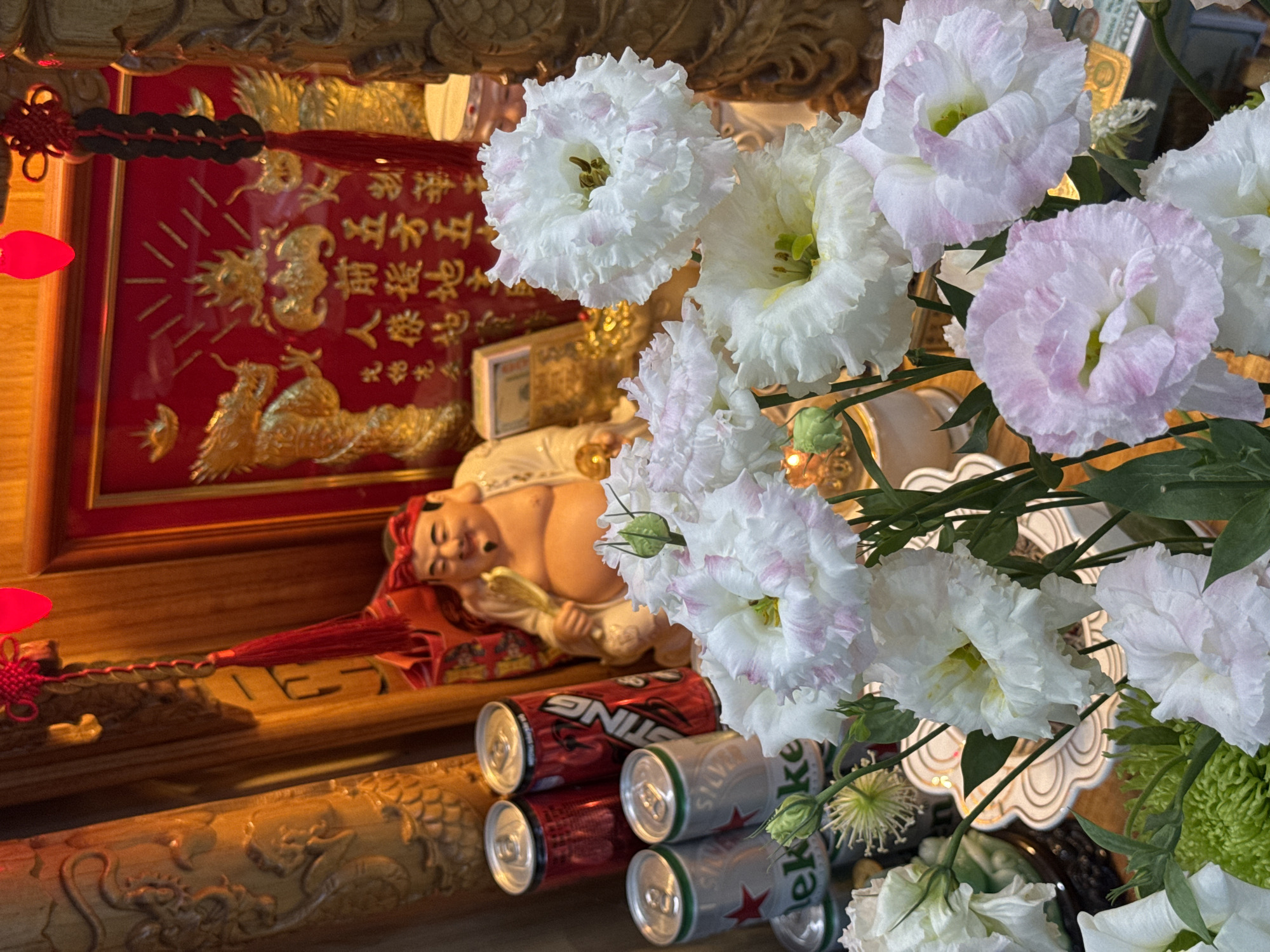 The image depicts an altar or shrine, likely in a home or religious establishment in Nam Nha Trang Ward, Vietnam. In the background, a golden dragon mural adorns a red backdrop, framed by ornate wooden carvings. A jovial Buddha statue sits to the right, holding a fan. In front of these elements, a collection of white and pink lisianthus flowers are arranged in a vase, some in full bloom and others as buds. Several beverage cans are stacked behind the flowers, including red cans with "STING" branding and silver cans with the Heineken logo. The overall scene suggests a religious offering or a devotional space, possibly for a traditional Vietnamese celebration or everyday worship.