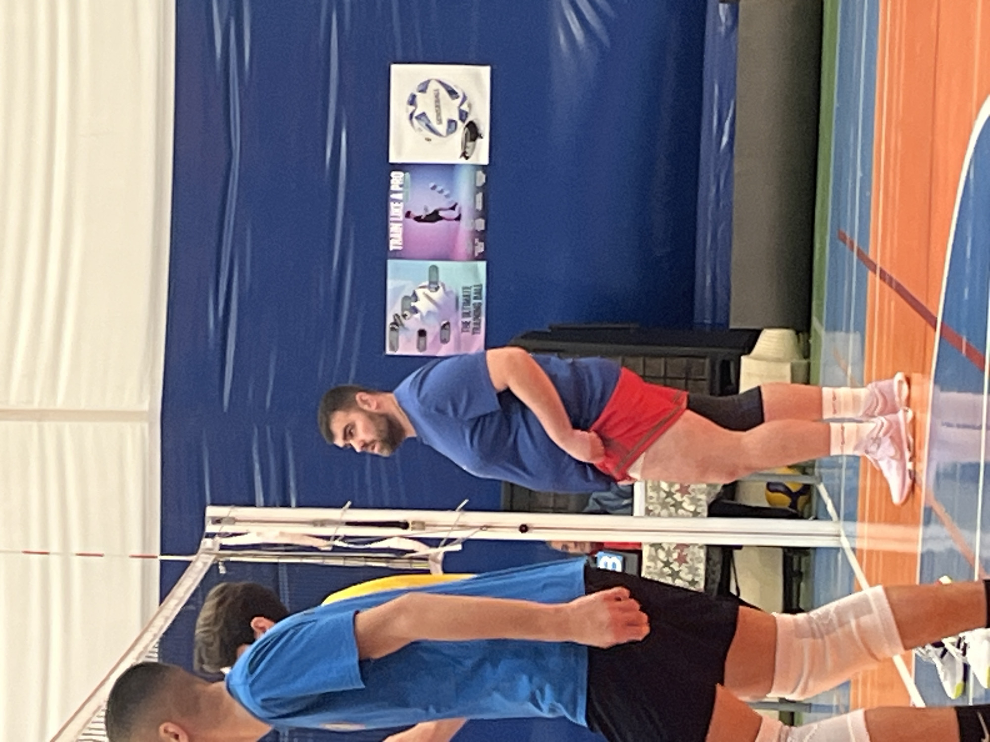 Two men in athletic wear are positioned near a volleyball net inside a gymnasium with blue wall coverings, an orange and blue court, and wall-mounted informational posters. The location is likely in Sofia, Bulgaria.