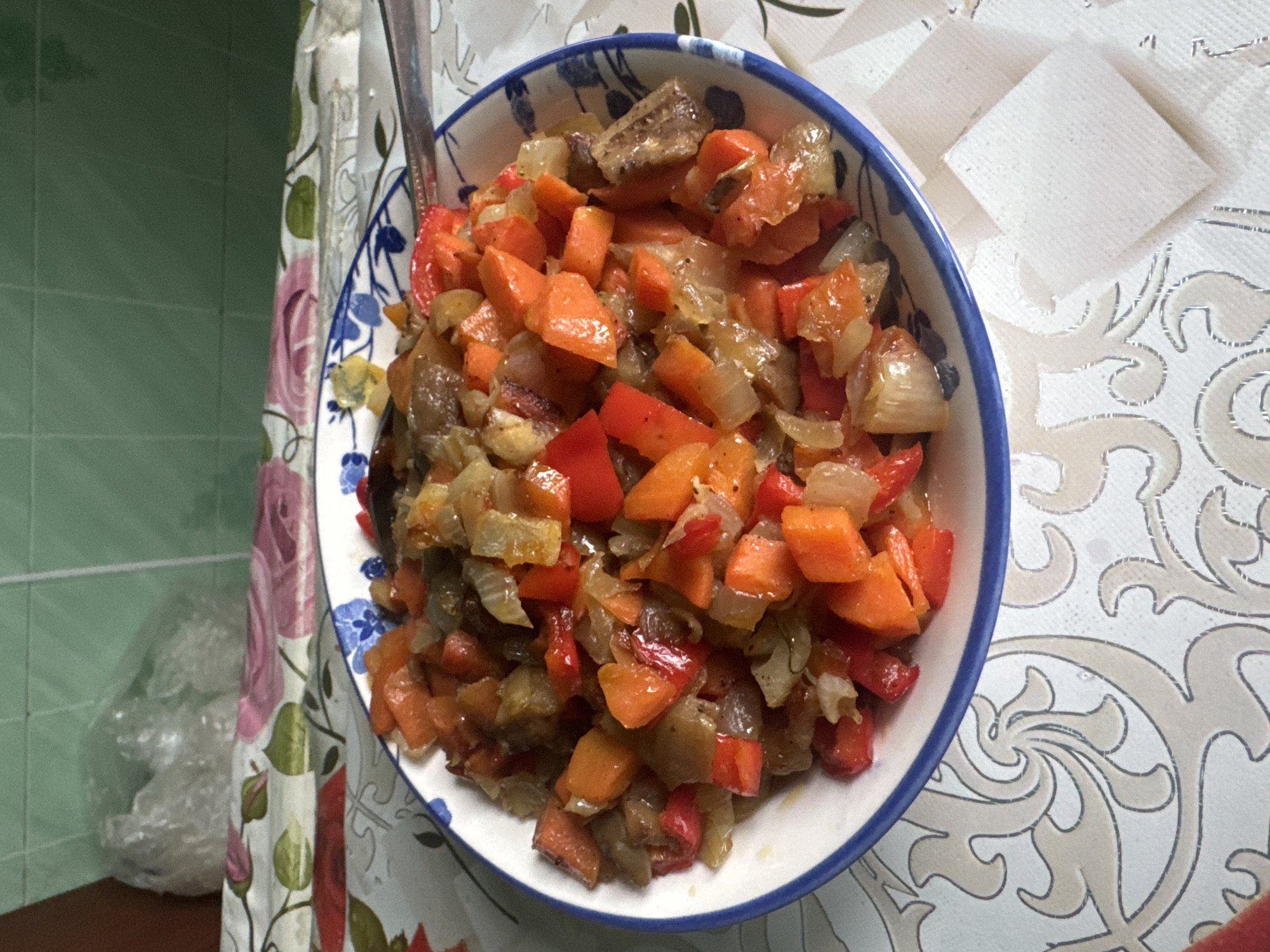 A close-up, overhead shot shows a bowl of vegetable stew, likely ratatouille, placed on a patterned tablecloth. The stew contains diced carrots, red bell peppers, and onions, all glistening with oil and seasoning. A spoon rests in the bowl, suggesting the meal is ready to be eaten. The tablecloth features an intricate floral and scrolling pattern in beige and white. In the background, green tiled walls and a plastic-wrapped item are visible. The scene appears to be set in a kitchen or dining area.