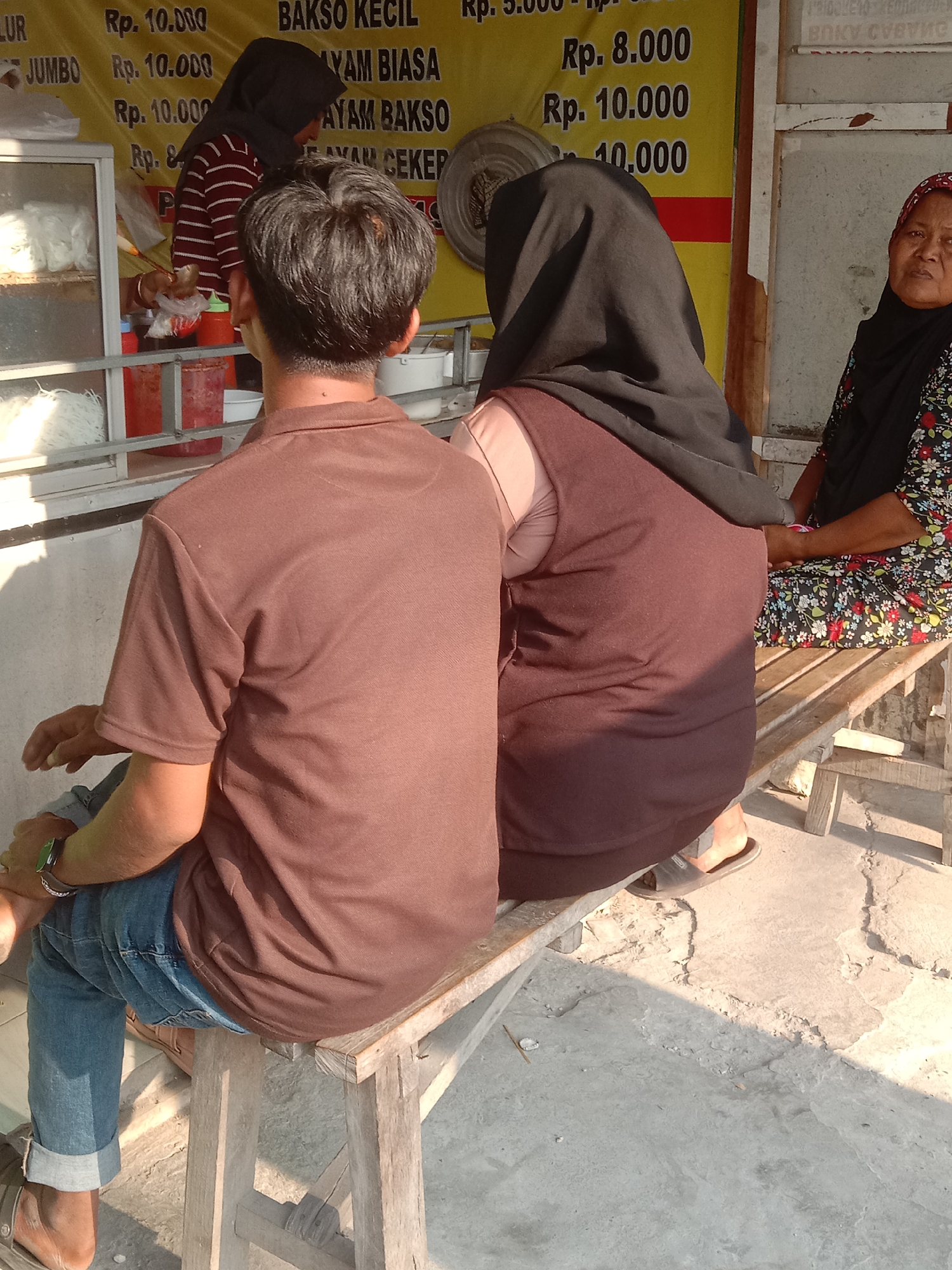 At an outdoor food stall in Sumberharjo, Indonesia, three individuals are visible. In the foreground, a man with dark hair, wearing a brown polo shirt and blue jeans, sits on a wooden bench. He is viewed from the back. To his right, a person wearing a black hijab and a dark brown vest over a light pink shirt sits on the same bench. Further to the right, a woman wearing a black hijab and a floral-patterned dress sits on the bench, looking towards the left. Behind the people, a yellow sign displays prices in Indonesian Rupiah. A glass display case filled with white noodles is visible to the left. The ground appears to be concrete, with visible cracks and debris. Sunlight casts shadows across the scene.