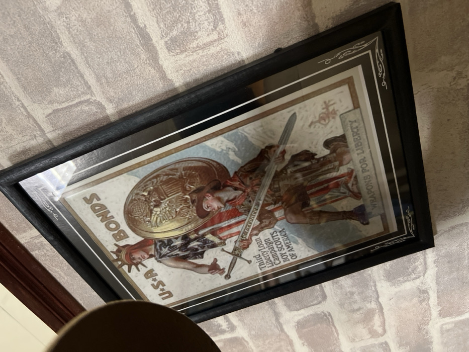 A framed vintage poster displays a patriotic scene. "U.S.A Bonds" is prominent, next to an image of the Statue of Liberty. Another figure, possibly a soldier or scout, wields a sword and shield. Text on the poster reads "Third Liberty Loan Campaign Boy Scouts of America" and "Weapons for Liberty." The poster's design includes elaborate embellishments, captured within a dark, ornate frame. The artwork is mounted on a brick wall.