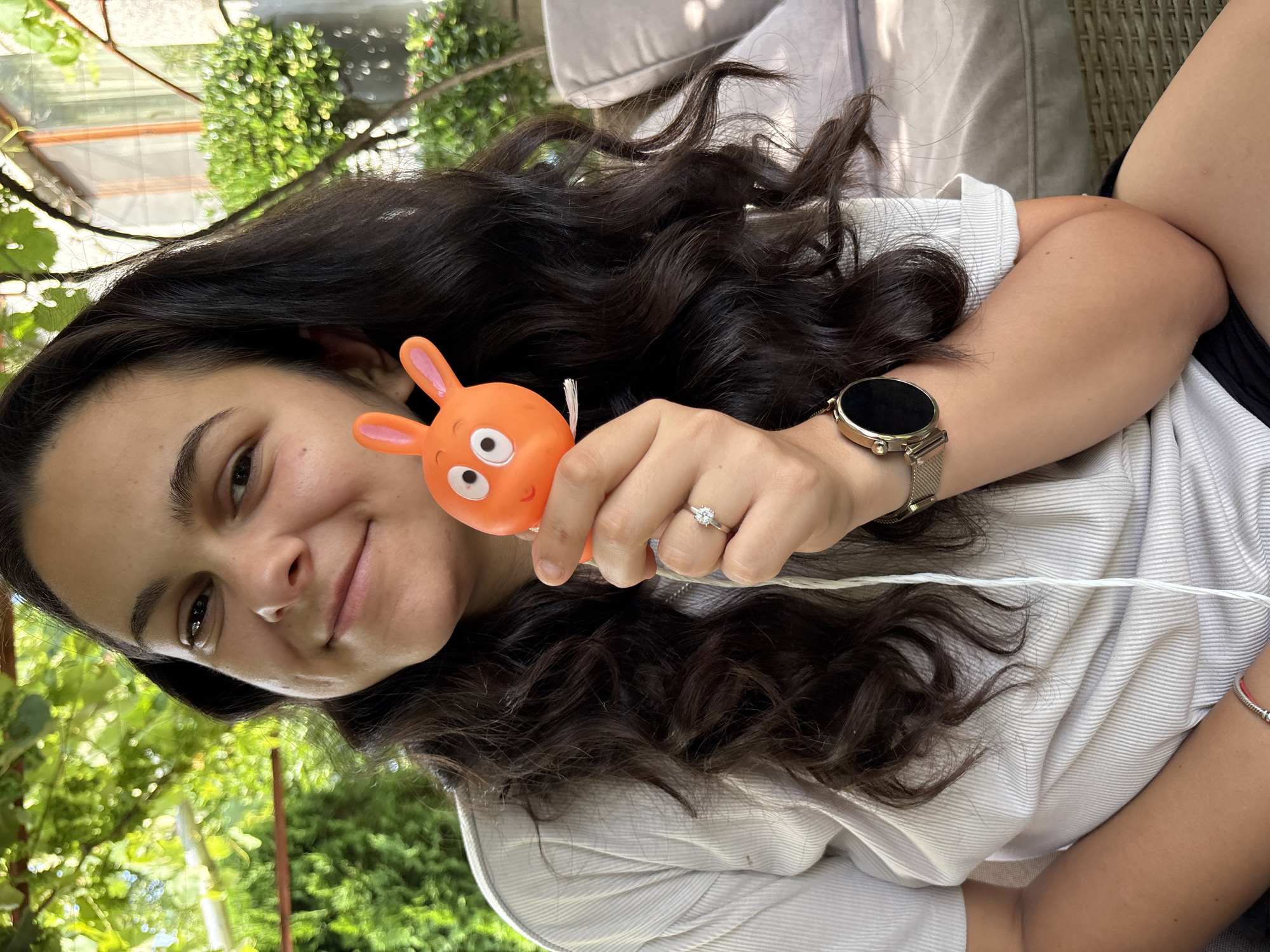 In this close-up, vertically oriented outdoor shot taken in Oreshak, Bulgaria, a young woman with long, dark, wavy hair is playfully holding an orange, rabbit-shaped toy with big, round eyes. She is wearing a light-colored, subtly striped t-shirt and a silver watch on her left wrist. A delicate ring with a diamond is visible on her right ring finger. Her expression is a cheerful smile, suggesting a lighthearted moment.
The background is softly blurred, but hints of lush green foliage and what appears to be an outdoor seating area with woven furniture and a cushion suggest a relaxed, possibly backyard or garden setting. Sunlight filters through the leaves, indicating it's daytime. A thin white string is visible running across the frame from the toy towards the woman's chest. There is no visible text in the image. The overall mood is one of joy and casual enjoyment.