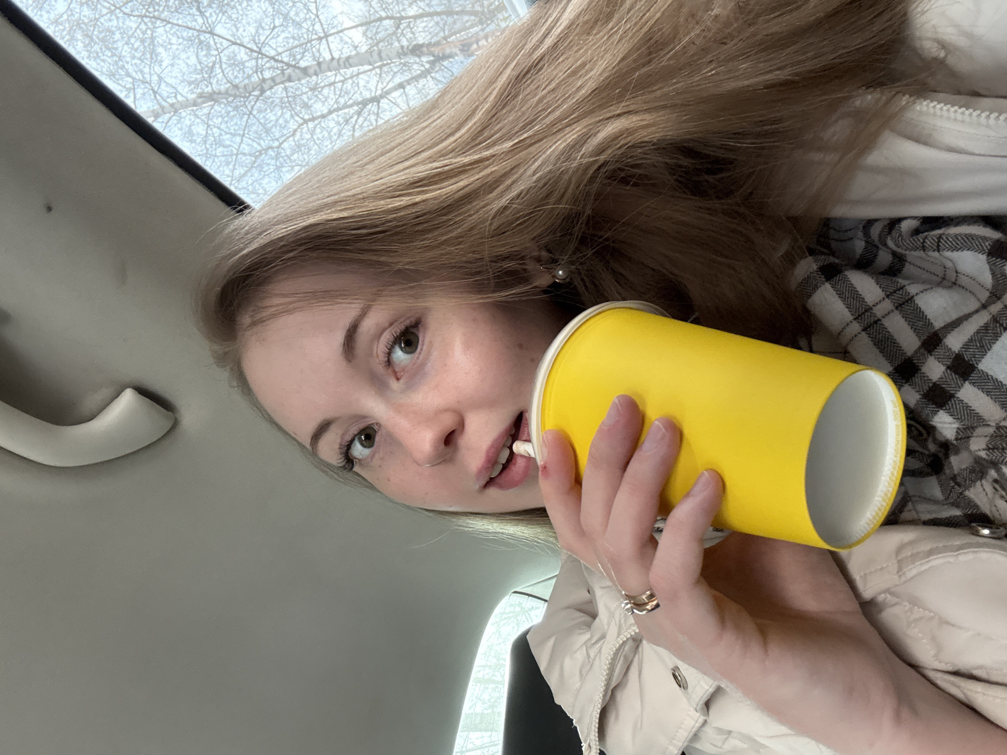 A young, light-skinned woman with long, blonde hair is holding a yellow cup with a white interior in front of her face. She has light eyes and fair skin. The woman is inside a car, as evidenced by the car's interior features visible in the background, including the ceiling, a grab handle, and a view of the sky and trees outside the window. She is wearing a beige puffer jacket over a black and white checkered shirt. She appears to be drinking from a straw. The location is unknown.