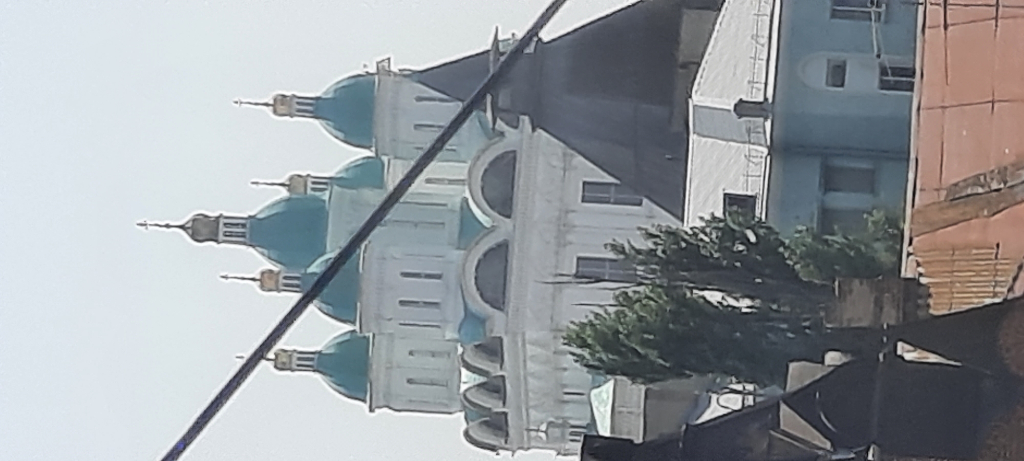 This image, captured from a distance and partially obscured by wires and foreground structures, depicts the Assumption Cathedral in Astrakhan, Russia. The prominent architectural feature is the cathedral's distinctive light blue domes, topped with gold crosses. Several of these domes are visible, arranged in a cluster around a central, taller dome. The cathedral itself appears to be a large, white structure with multiple levels and arched windows.
In the middle ground, several trees with dense green foliage stand in front of the cathedral. To the right of the trees and partially in front of the cathedral's lower levels, is a dark, conical roof structure, possibly part of another building or an annex.
In the foreground, the roofs of several buildings are visible. These range from dark, angular structures to a lighter-colored roof with a patterned surface, and a reddish-brown tiled roof. A light blue building with windows is visible behind the trees and the conical structure.
The sky is overcast or hazy, with a pale, washed-out appearance. The overall impression is of a cityscape view featuring a significant religious landmark.