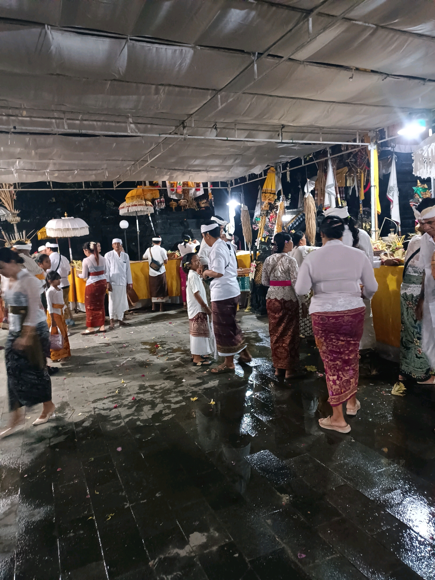 This image depicts an outdoor gathering, possibly a religious ceremony or festival, taking place at night under a white tent-like covering. The ground is wet and reflective, suggesting recent rain or a damp environment, with scattered flower petals on the pavement. Several individuals are present, many wearing traditional attire including sarongs and white shirts or blouses.
Specifically, the scene appears to be located in Kediri, Indonesia, within a temple or ceremonial ground. The attendees are a mix of adults and children. In the foreground, a person in a dark patterned skirt and light-colored shoes walks on the wet ground, their movement blurred. To the right, a woman in a vibrant purple patterned sarong and beige sandals stands, facing away from the camera.
Further into the scene, a group of people are gathered around a yellow-draped table or altar. Two white umbrellas, adorned with intricate patterns, are prominent. Several individuals in white traditional clothing, including head coverings, are visible, some interacting with each other. The background is dark, with some foliage visible beyond the tent. Flags and decorative elements are hung from poles or structures, adding to the festive or ceremonial atmosphere. The lighting is a mix of ambient and artificial lights, creating some highlights and shadows. The overall impression is one of a community event in progress.