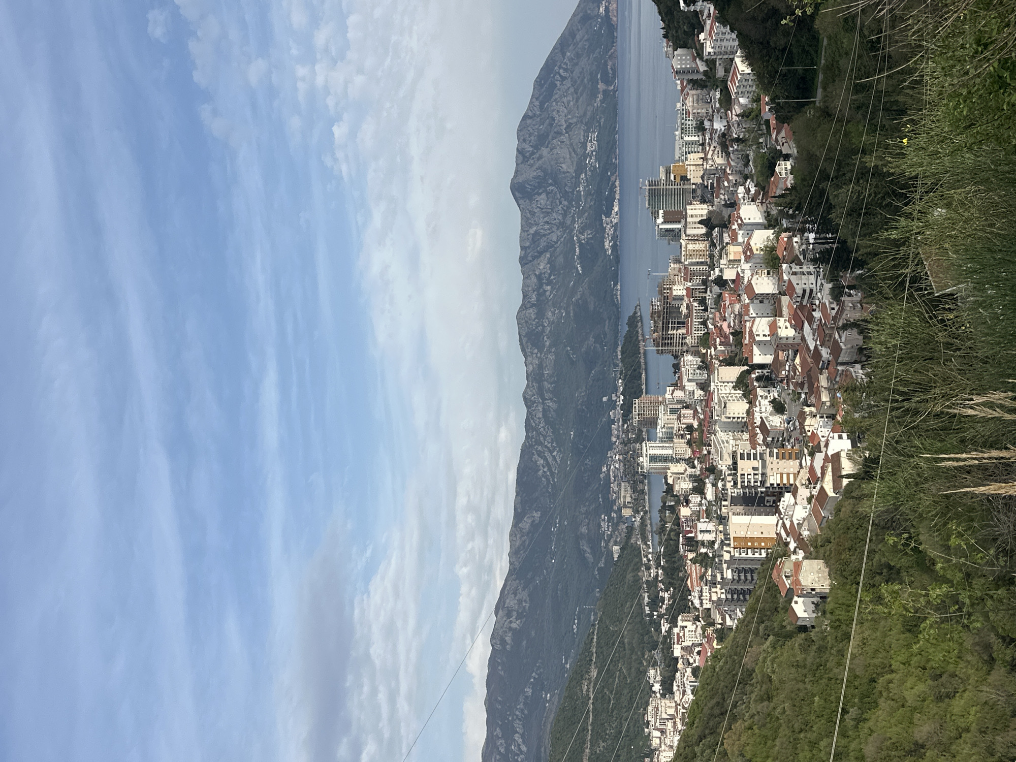 The image shows an elevated view of Budva, Montenegro. The scene includes a mountain range with a shoreline on one side and the city of Budva on the other. The sky is visible. The buildings are mostly white and red. Green vegetation is seen at the right edge of the image. Power lines are visible in the foreground.