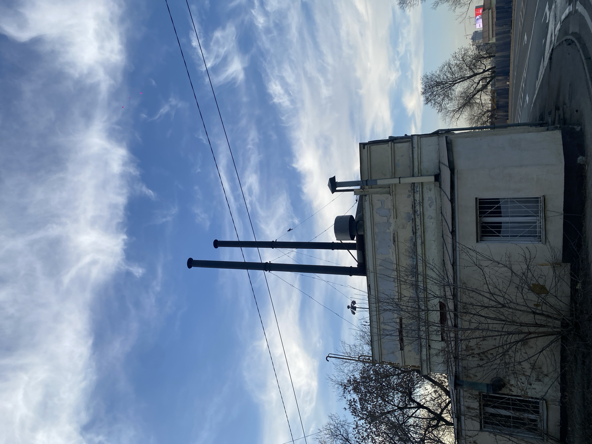 The image depicts an exterior scene in Khimki, Russia, focusing on an aged utilitarian building under a partly cloudy sky. The sky is bright blue with scattered white, wispy clouds. Three small, faint pink dots are visible high in the sky. Several dark electrical wires traverse the upper frame.
The central subject is a two-story light beige or off-white building with peeling paint and discolored sections, indicative of wear. Its windows are secured with vertical and horizontal metal bars. The roof features two prominent tall, dark metal exhaust stacks, stabilized by guy wires anchored to the roof structure. Also on the roof are a smaller dark cylindrical tank, another smaller dark vent with a square cap, and a multi-pronged antenna-like device. Various external pipes and conduits are visible on the building's facade.
To the left of the building, a bare deciduous tree with a dense network of branches partially obstructs the view of the building's side. Another bare tree is visible further in the background. To the right of the building, an asphalt road with white lane markings (including arrows) curves away. A dark, corrugated metal fence runs parallel to the road in the mid-ground. In the distant background, several tall structures with visible red-lit panels or screens can be observed, potentially indicating urban development or commercial signage. No individuals are present in the scene, and no explicit actions or interactions are depicted.