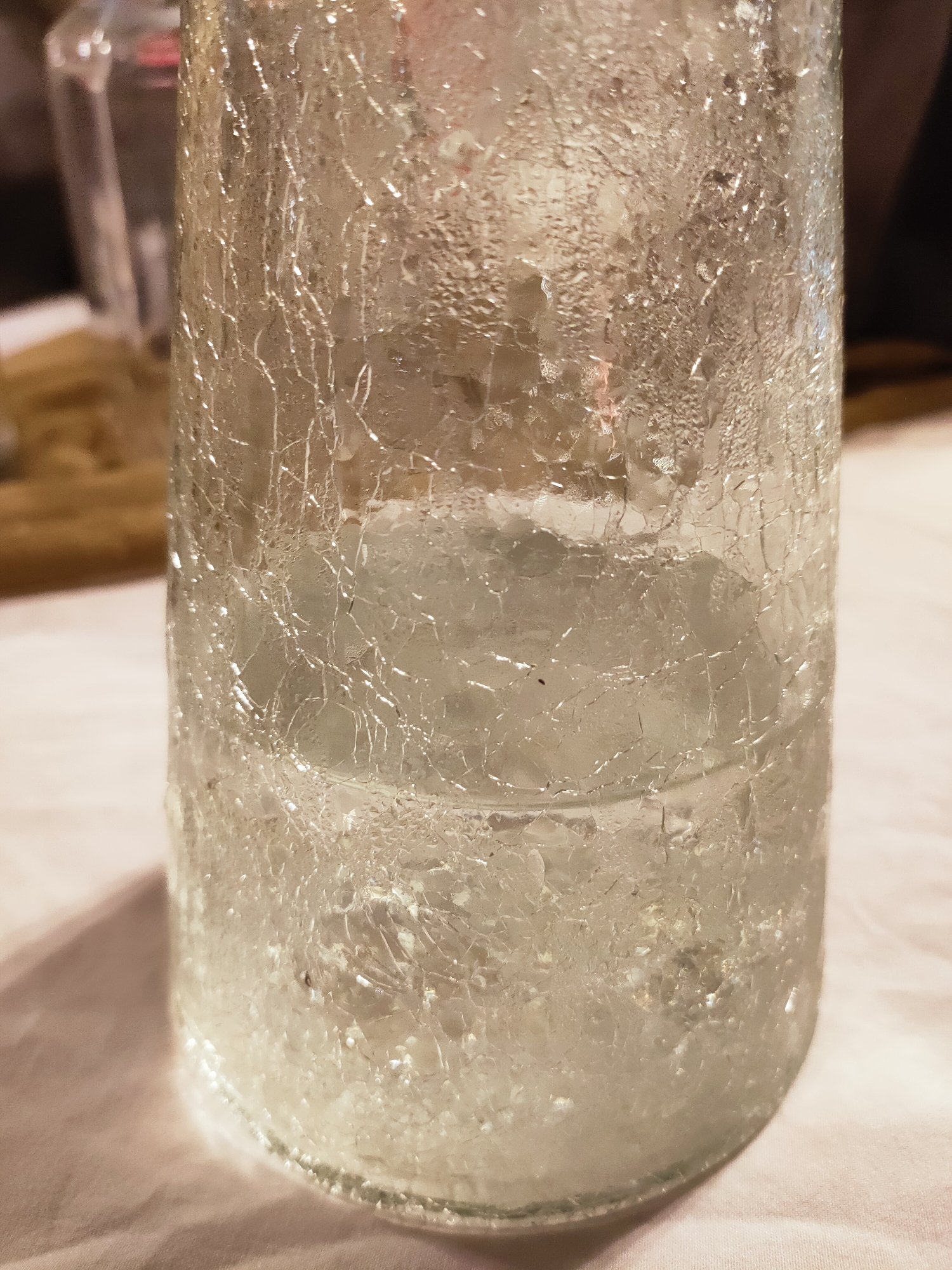 The image shows a close-up of a glass container filled with ice. The glass has a textured, crackled surface. The ice inside is broken into small pieces and appears to be melting. The background is out of focus, but suggests an indoor setting with a tablecloth and other objects. There is no information about the city of Cebalat, Tunisia present in the image.