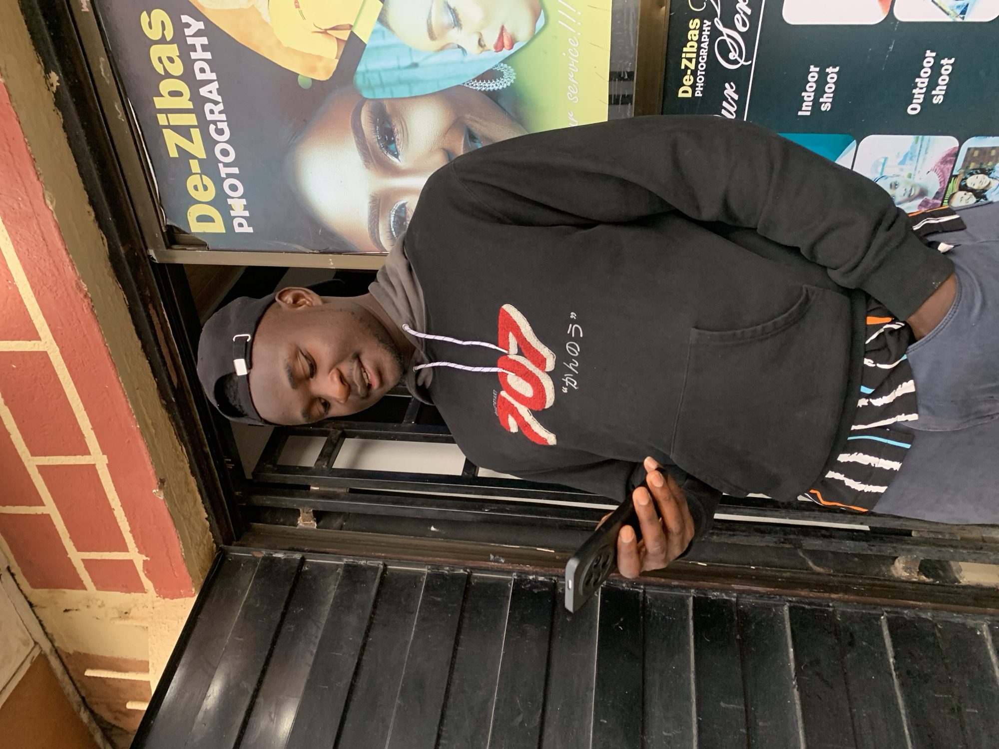 A Black man wearing a black hoodie with "707" in red and white lettering, a black baseball cap, and grey pants stands in front of a shop. He is looking down at his phone, which he holds in his right hand. The shop has a black shuttered door to his left and a large poster for "De-Zibas Photography" to his right. The poster features images of women and text advertising photography services. The background includes a red brick wall above the shop entrance. The scene appears to be located in Jos, Nigeria, suggested by the presence of local business signage.