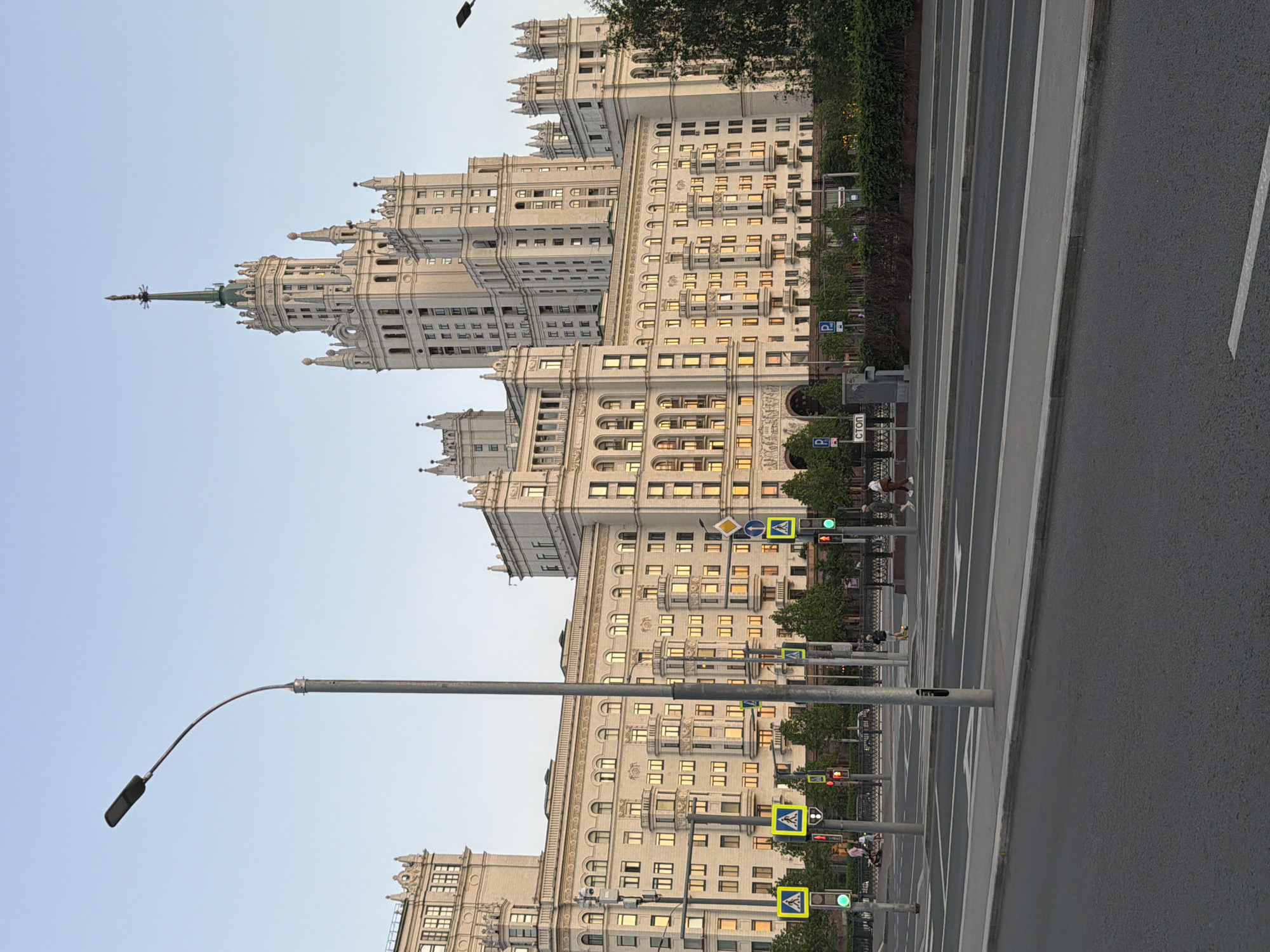 The image depicts the Kotelnicheskaya Embankment Building, a Stalinist skyscraper in Moscow, Russia. The building is prominent in the background, with its distinctive architectural style and spire. In the foreground, there is a road with multiple lanes and a pedestrian walkway. There are traffic lights and pedestrian crossing signs visible. The scene appears to be taken during daylight hours, with soft lighting. There are trees and foliage along the road. The image is taken from a ground-level perspective. There are a few people walking in the background.