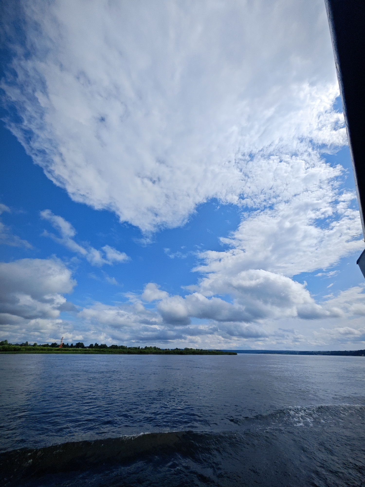 The image depicts a wide, dark blue river under a vast sky filled with white, fluffy clouds. The water shows a subtle ripple pattern, and a darker wake is visible in the foreground, suggesting movement. On the distant horizon, a line of green trees forms the bank of the river, with a faint orange structure, possibly a crane, visible among them. The sky is a bright blue, with a strong contrast between the blue sky and the white clouds, some of which are dense and others wispy. The scene is captured from a perspective near the water's surface, possibly from a boat, as indicated by the dark, out-of-focus object on the far right edge of the frame. The overall impression is one of a calm, natural landscape under a dynamic sky. There are no people or animals visible in the image. The location in the city of Krutoy Log, Russia is a riverbank scene.