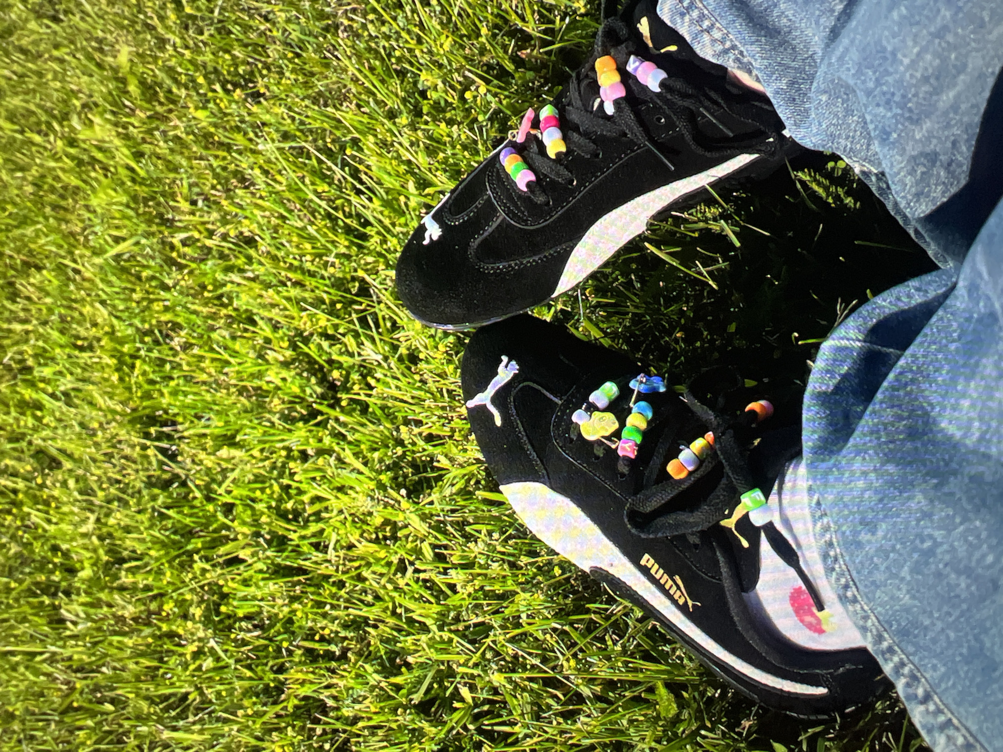 The image shows a pair of black Puma sneakers adorned with colorful beaded lace charms, resting on a lush green lawn. The laces are threaded with beads of various colors, including pink, yellow, orange, green, and blue, creating a playful and personalized look. One shoe has a white Puma logo and a white stripe detail, while the other shoe is partially obscured. The person wearing the shoes is also wearing blue denim jeans. The sunlight casts shadows on the grass, indicating it is daytime. The overall scene suggests a casual outdoor setting. The location is not specified beyond "Unknown, Russia."
