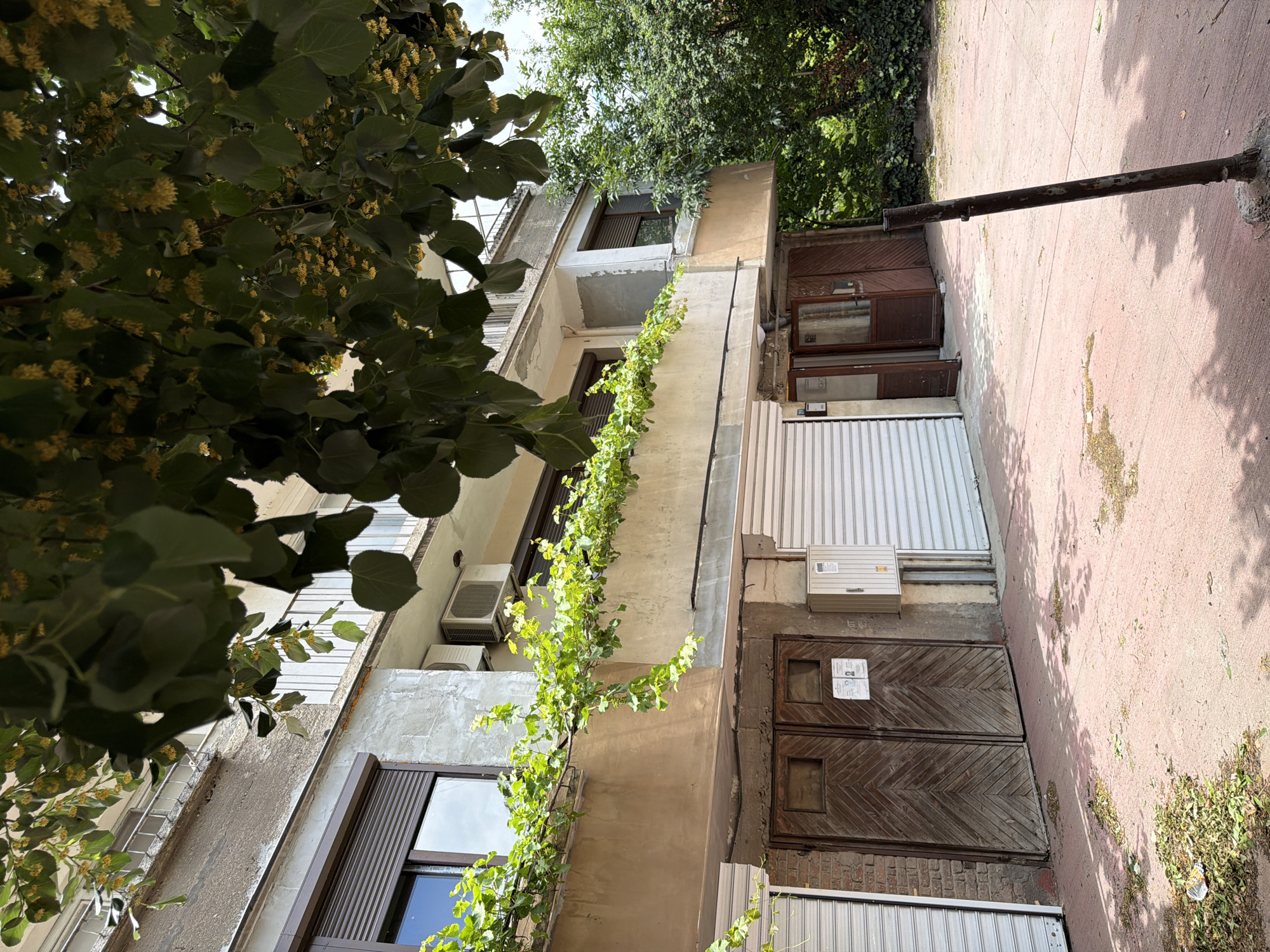 The image shows the ground floor and first floor of a concrete building with a beige-colored facade. The ground floor features two large, diagonal wooden doors and a white roller shutter. A smaller wooden door is ajar to the right of the wooden doors. Above the ground floor, on the first floor, a balcony is visible with a railing and a vine growing along its edge. Two air conditioning units are mounted on the building's facade. In front of the building, a red-colored pavement is visible, with scattered green leaves and some debris. A dark metal pole stands in the foreground, tilted. Lush green trees and foliage are present to the right of the building. The scene appears to be located in an urban environment, possibly a residential or commercial area.