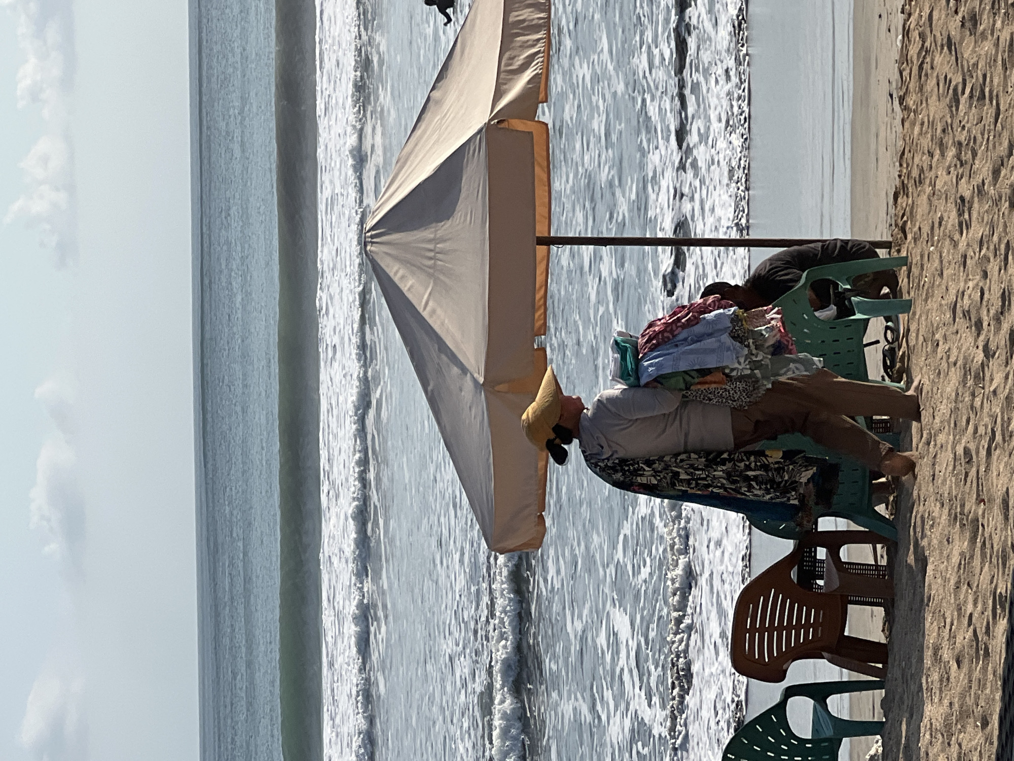 A person wearing a wide-brimmed hat and carrying a load of colorful textiles stands on the beach in Kuta, Indonesia, in front of a large, light-colored umbrella. Several plastic chairs, one brown and two green, are arranged on the sand. The ocean with gentle waves is visible in the background, with a lone surfer in the distance. The scene appears to be during daylight hours with a clear sky.