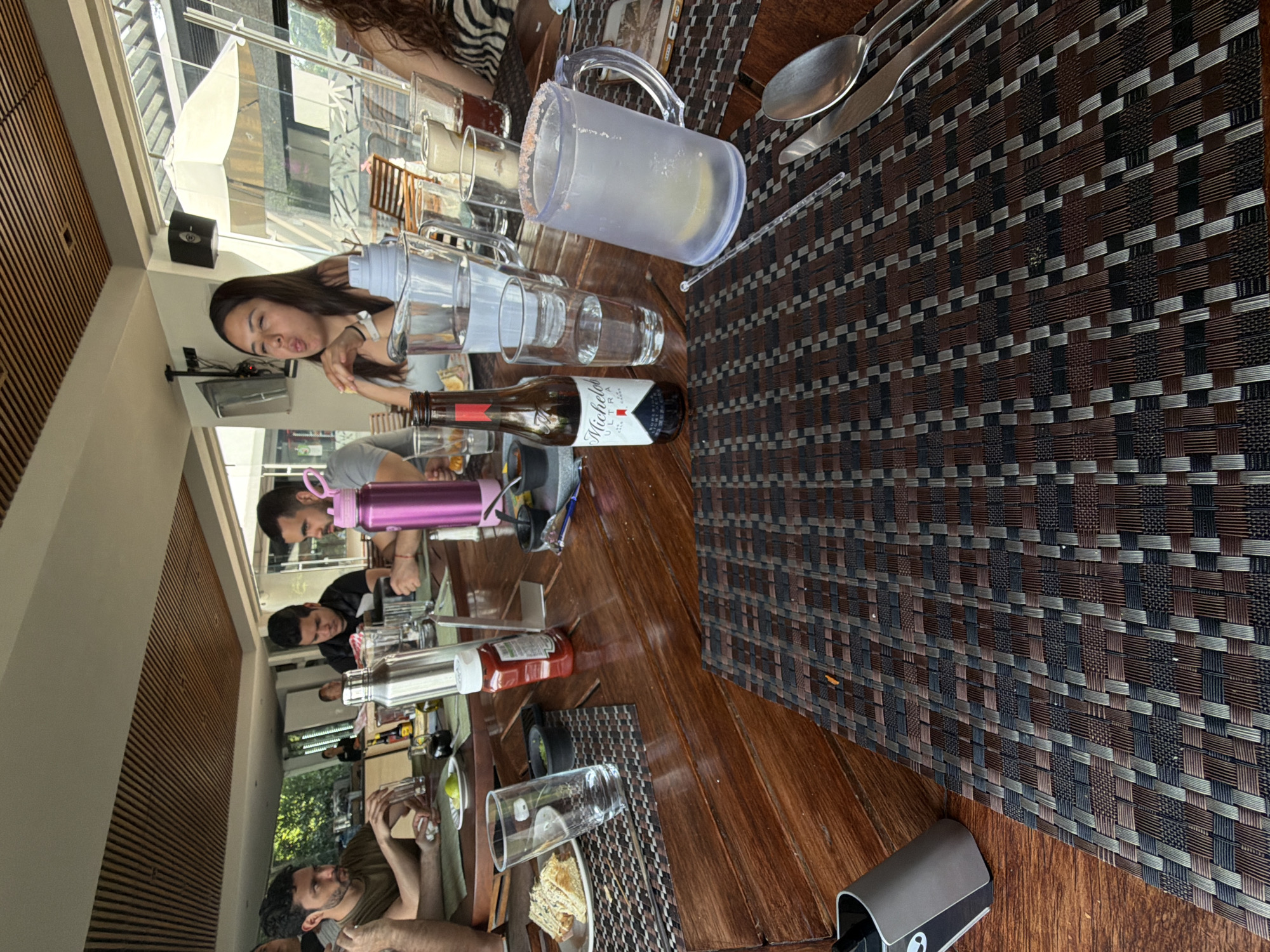 The scene captures a group of people gathered around a wooden table, potentially in a restaurant or bar, suggesting a social setting. The location is Puebla, Mexico.
Several individuals are visible around the table; one woman is in the foreground, while others are seen further down the table. Some of them are engaged in conversation or observing something on the table.
Numerous objects are spread across the table, including a bottle of Michelob Ultra beer, several glasses, a pitcher with a salted rim (likely for micheladas), cutlery, and a bottle of ketchup. There are also plates with food items, indicating that the group is eating or has eaten.
The table is covered with a patterned placemat, and the environment seems to be indoors, possibly a restaurant with a view, indicated by the presence of windows or an open area. The setting suggests a casual or informal gathering.