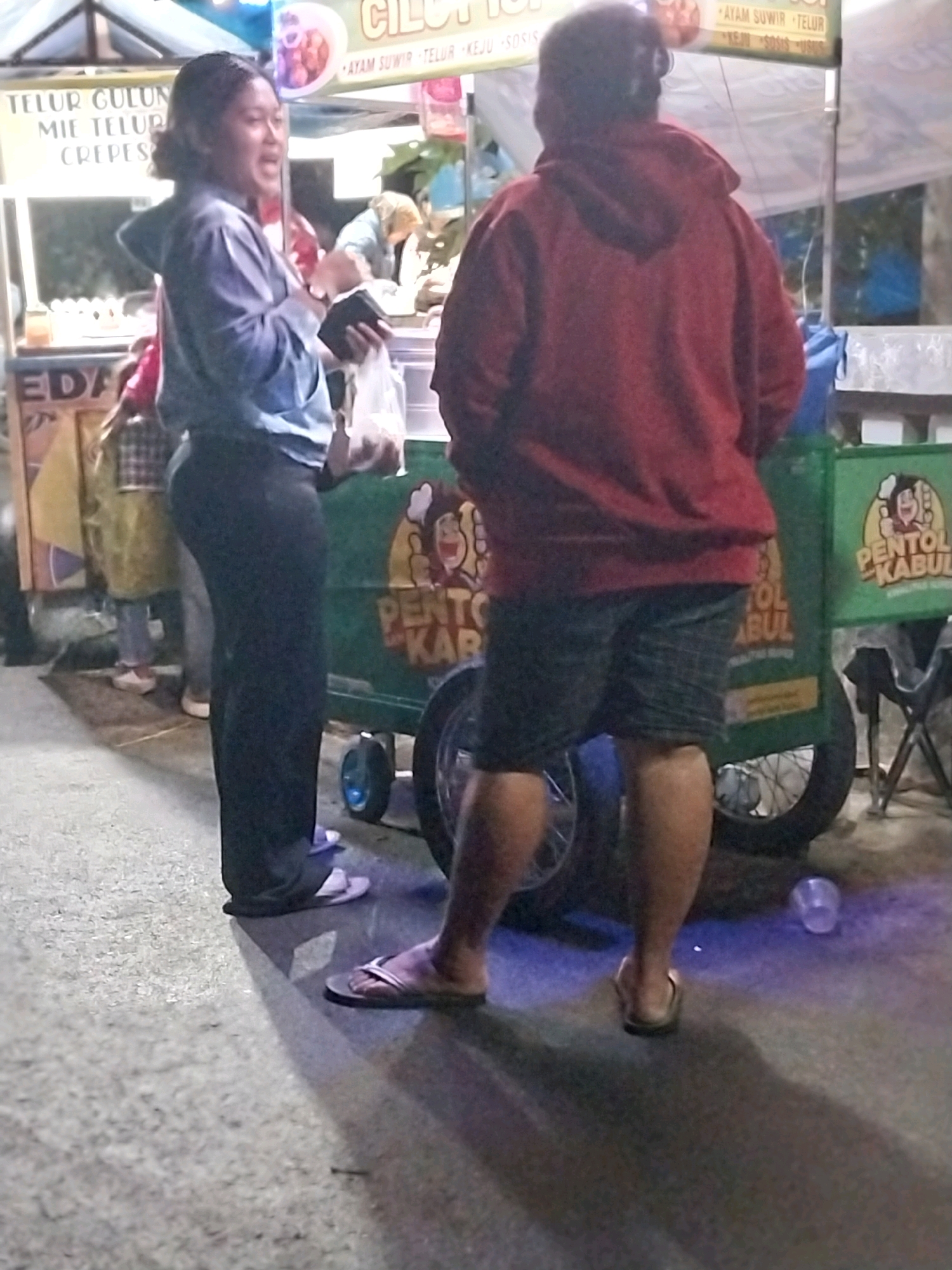 In the city of Nglegok, Indonesia, a street food vendor stall is depicted at night. The stall features a green cart with yellow signage that reads "PENTOL KABUL." A woman in a light blue long-sleeved shirt and black pants stands to the left of the cart, holding a black wallet and a white bag. She is facing towards the right, with her mouth open as if speaking. To her right stands a person wearing a red hooded sweatshirt and plaid shorts. This person is facing away from the camera and appears to be interacting with the woman. The ground is paved and dimly lit, with shadows cast by the individuals and the stall. The background shows glimpses of other stalls and tents, indicating a busy marketplace.