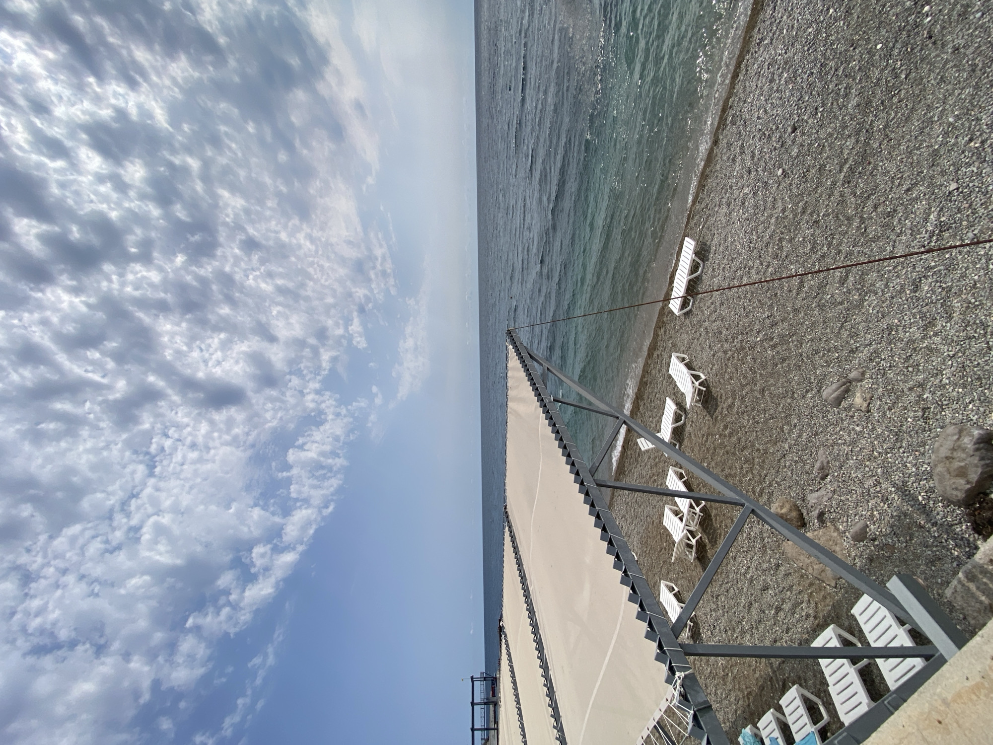 The image depicts a seaside location, likely a beach in the Gasprinsky Settlement Council, Ukraine. The scene is dominated by the ocean, which has a clear, greenish-blue hue and gentle waves lapping onto a pebble beach. A row of empty white lounge chairs is arranged on the beach, some under a beige awning supported by a metal frame. The sky is partly cloudy, with a mix of white and grey clouds against a bright blue backdrop. In the distance, a dark structure with a platform is visible near the water's edge. The overall impression is one of a quiet, possibly off-season, beach resort. No people are visible in the image.