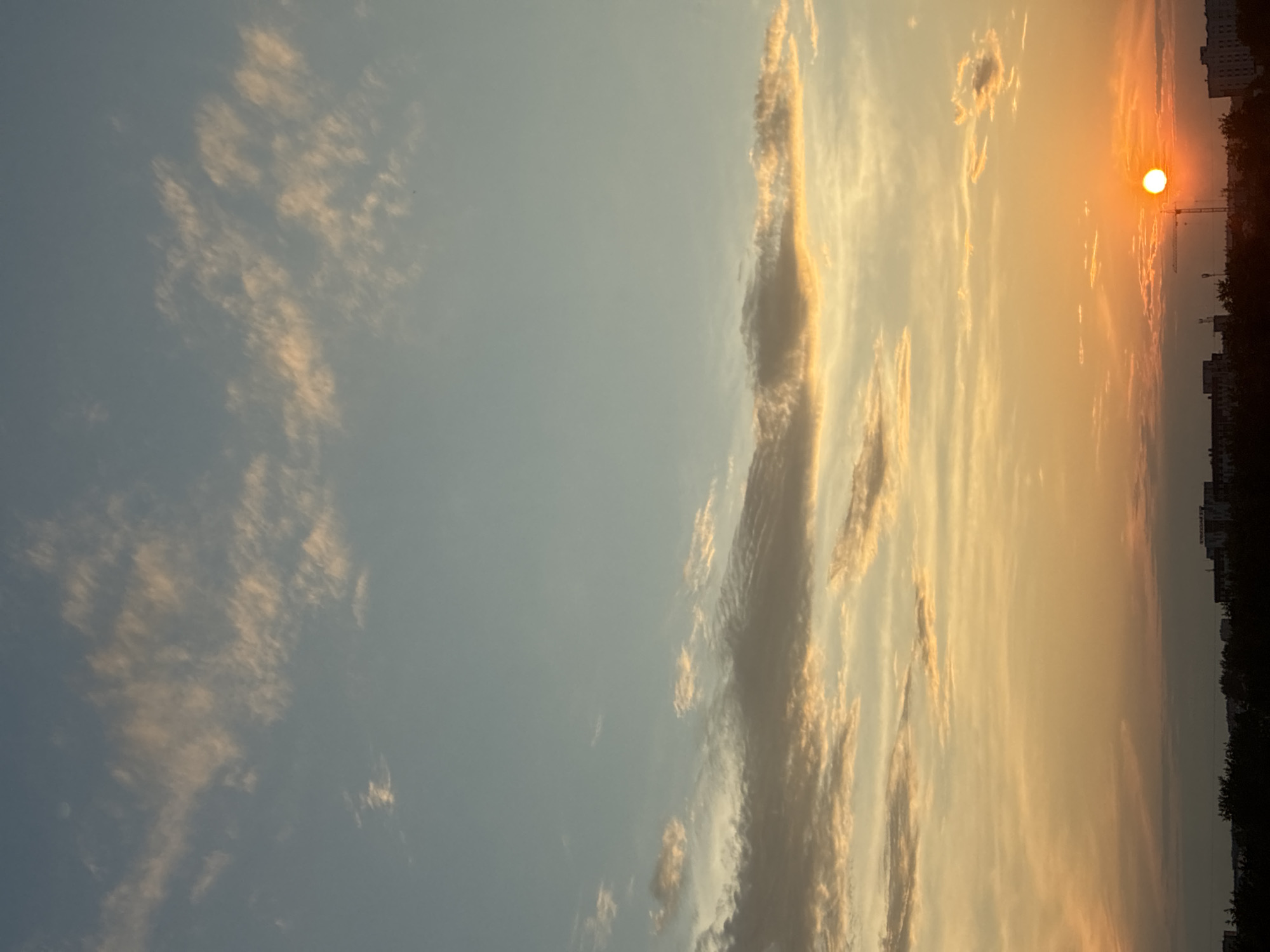 The image captures a sunset over an urban landscape. The sky transitions from a pale blue at the top to a vibrant orange and yellow near the horizon, with scattered wispy clouds catching the light. The sun, a bright orange orb, is visible near the right side of the frame, low on the horizon. Silhouetted against the sky is a dark line of trees and buildings, indicating a city or town. A construction crane is also visible near the sun. The overall impression is that of a peaceful evening scene.