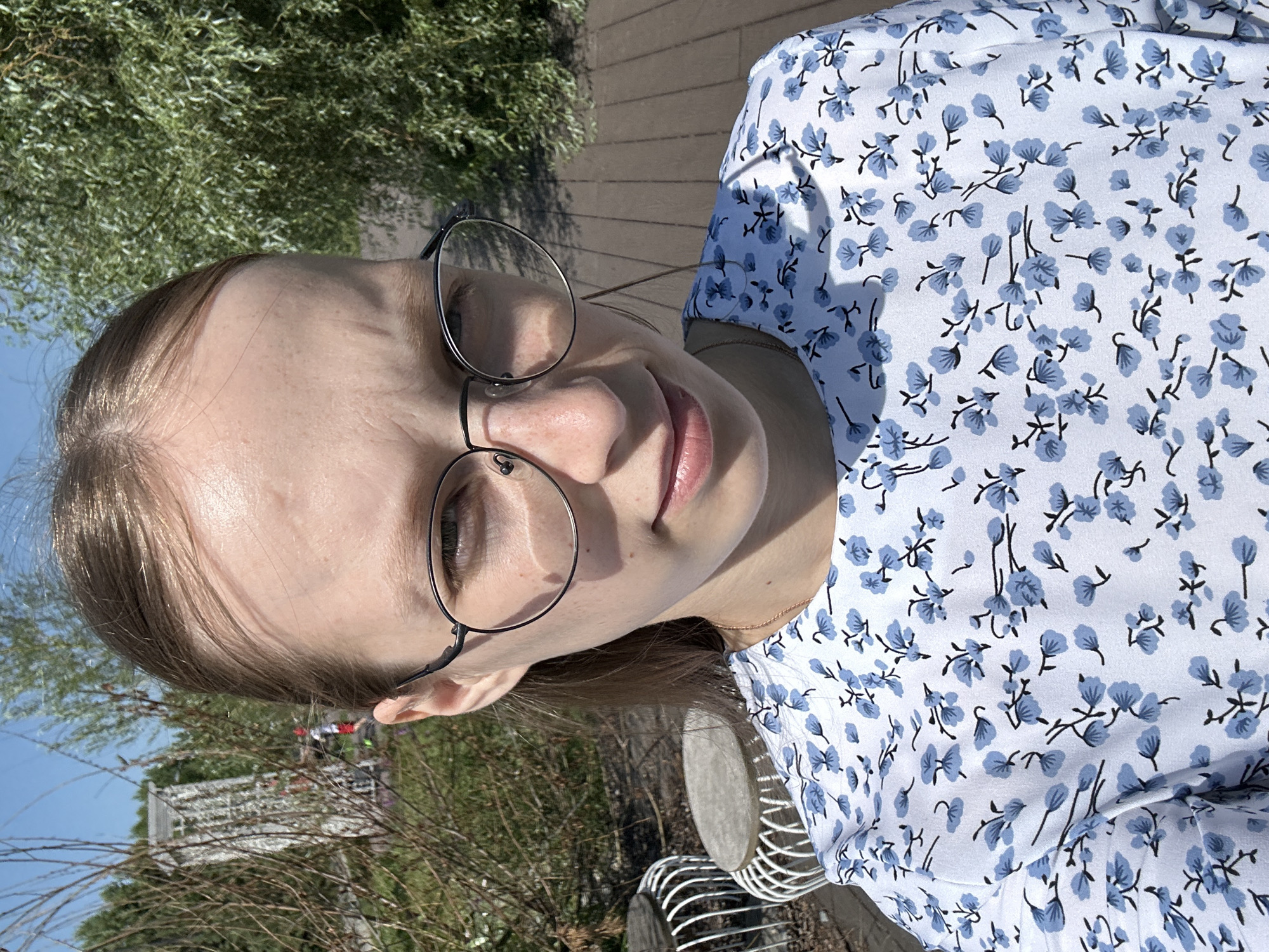A young woman with fair skin, brown hair, and glasses is taking a selfie outdoors. She is wearing a white dress with a blue floral pattern. The background suggests a park or outdoor seating area with wooden planks on the ground, some bushes, and trees. In the distance, a white structure, possibly a gazebo or trellis, is visible. The lighting indicates it is daytime, with a clear blue sky.