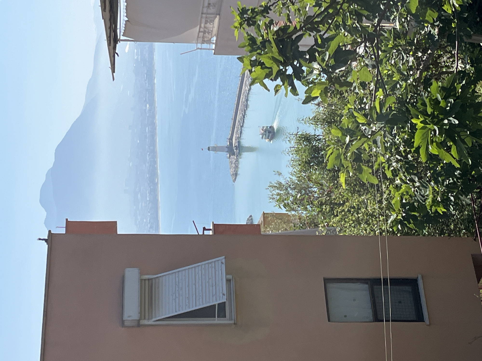 The image presents a bright and clear coastal landscape, likely capturing a view in Alanya, Turkey, from an elevated vantage point.
In the foreground, the top section of a light peach-colored building is visible. It features a window on the left with a partially opened white slatted shutter and a second, darker rectangular window to its right. Above and to the right of the building, vibrant green foliage from trees partially frames the scene, suggesting lush vegetation nearby.
The midground showcases a calm, turquoise-blue sea. A long, grey pier or breakwater extends horizontally into the water from the right side of the frame. At the far end of this structure stands a distinctive white lighthouse with a red top, from which a small, solid red flag, presumed to be the Turkish flag given the location context, flies. To the right of the lighthouse, a small boat with a dark roof is actively moving across the water, leaving a prominent white wake behind it.
In the background, a range of hazy, blue mountains stretches across the horizon under a clear, light blue sky. A faint outline of a coastal settlement can be discerned along the base of these distant mountains. The overall impression is one of a serene and sunny day at a scenic coastal location. There is no visible text in the image.