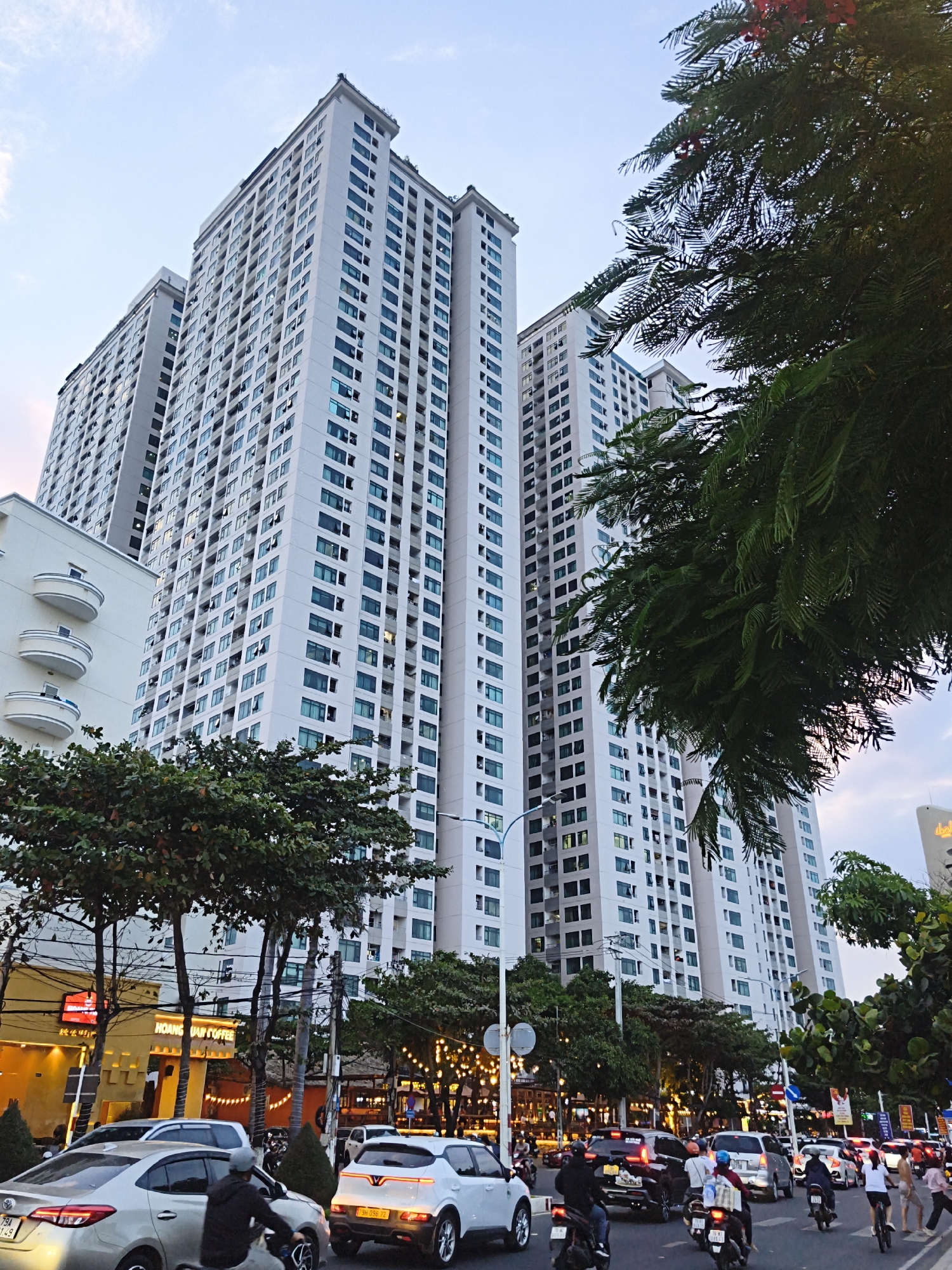 This media file depicts a street scene in Bac Nha Trang Ward, Vietnam, likely during the late afternoon or early evening. Towering, modern residential buildings dominate the background, their white facades punctuated by numerous windows. Some of these windows are illuminated, suggesting people are inside their homes as dusk approaches.
In the foreground, a busy street is filled with a mix of vehicles, including cars and motorcycles, indicating moderate traffic. People are seen riding the motorcycles, some carrying passengers or goods. Several shops and restaurants line the street, with one prominent establishment bearing yellow signage with Vietnamese text that appears to include "HOÀNG GIA COFFEE". Other businesses have string lights illuminating their facades, adding a festive atmosphere to the scene as daylight fades.
The sky above is a blend of soft blues and hints of pinkish-orange, characteristic of a sunset. Lush green trees with dense foliage are interspersed throughout the scene, some framing the view of the buildings and the sky. The overall impression is one of an active urban environment in a coastal city, with a blend of residential and commercial activity.