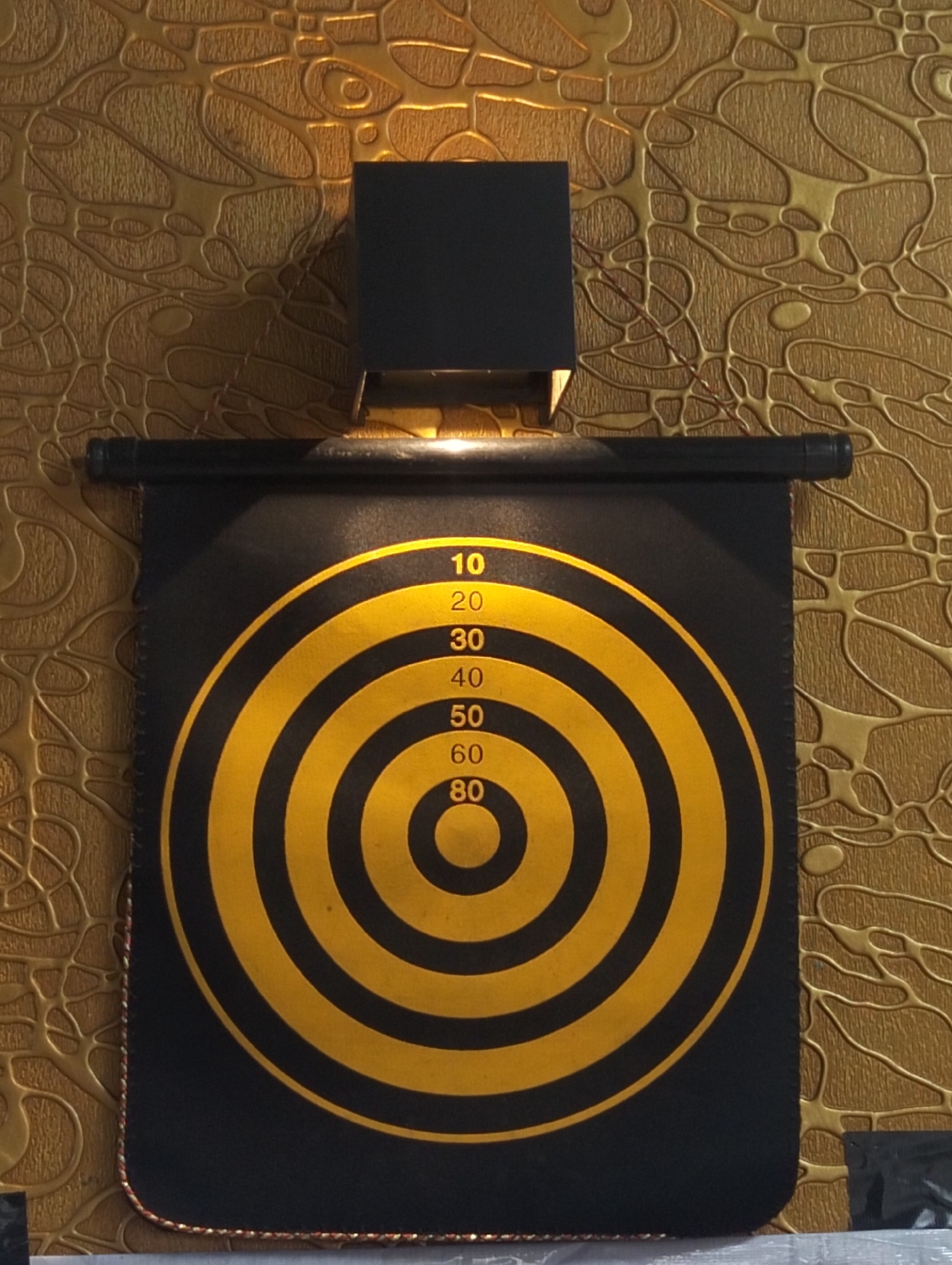 A dartboard hangs on a textured golden wall. The dartboard is black with concentric yellow rings and numerical targets. The numbers visible are 10, 20, 30, 40, 50, 60, and 80. A black light fixture is mounted above the dartboard.