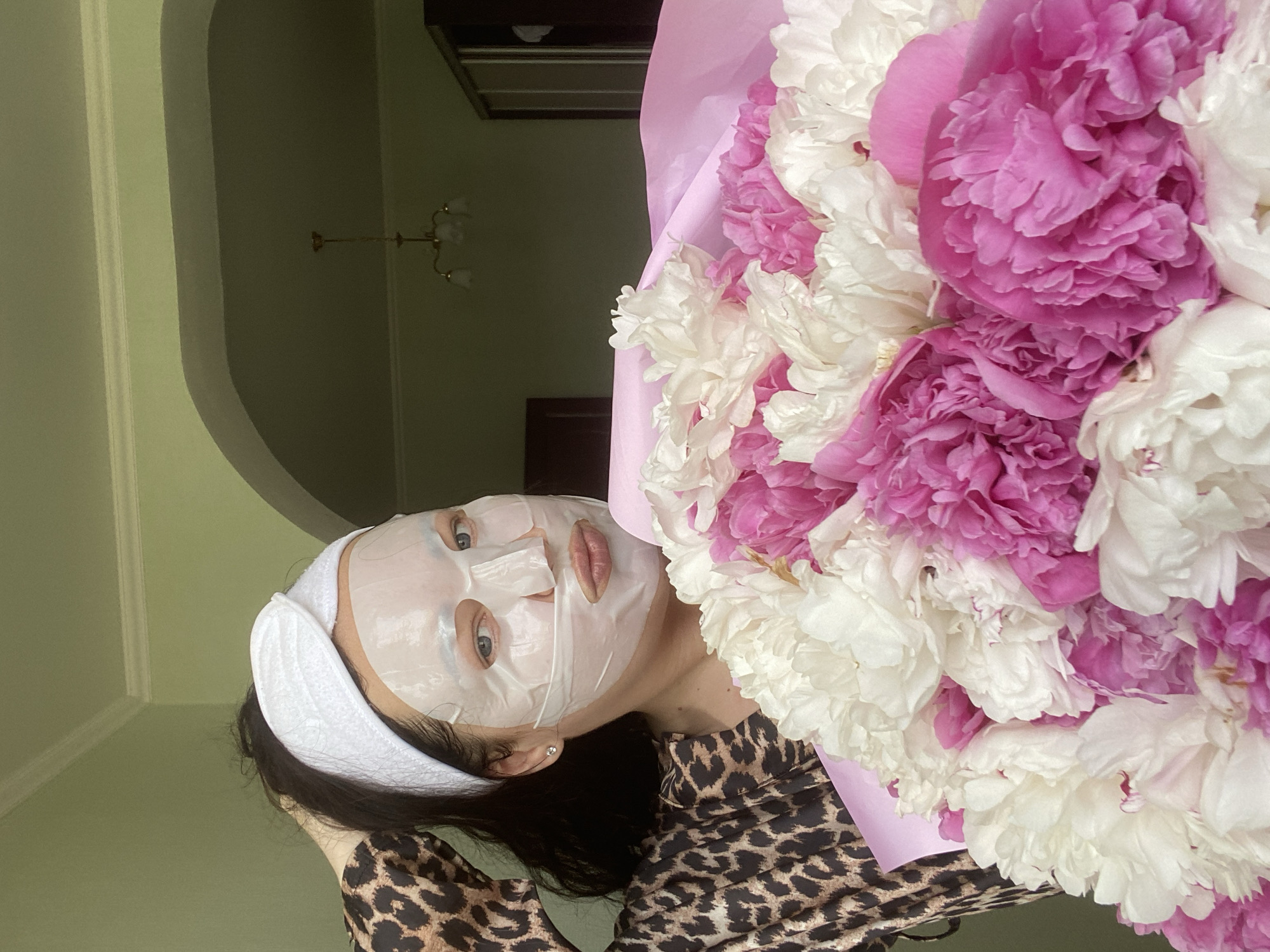 A woman wearing a face mask and a white headband is holding a large bouquet of pink and white peonies. She is wearing a leopard-print shirt. The background shows a room interior with a light green wall and an arched doorway. A chandelier is visible in the background. The location is most likely a private residence, but without additional information, the location in Sochi, Russia cannot be verified.