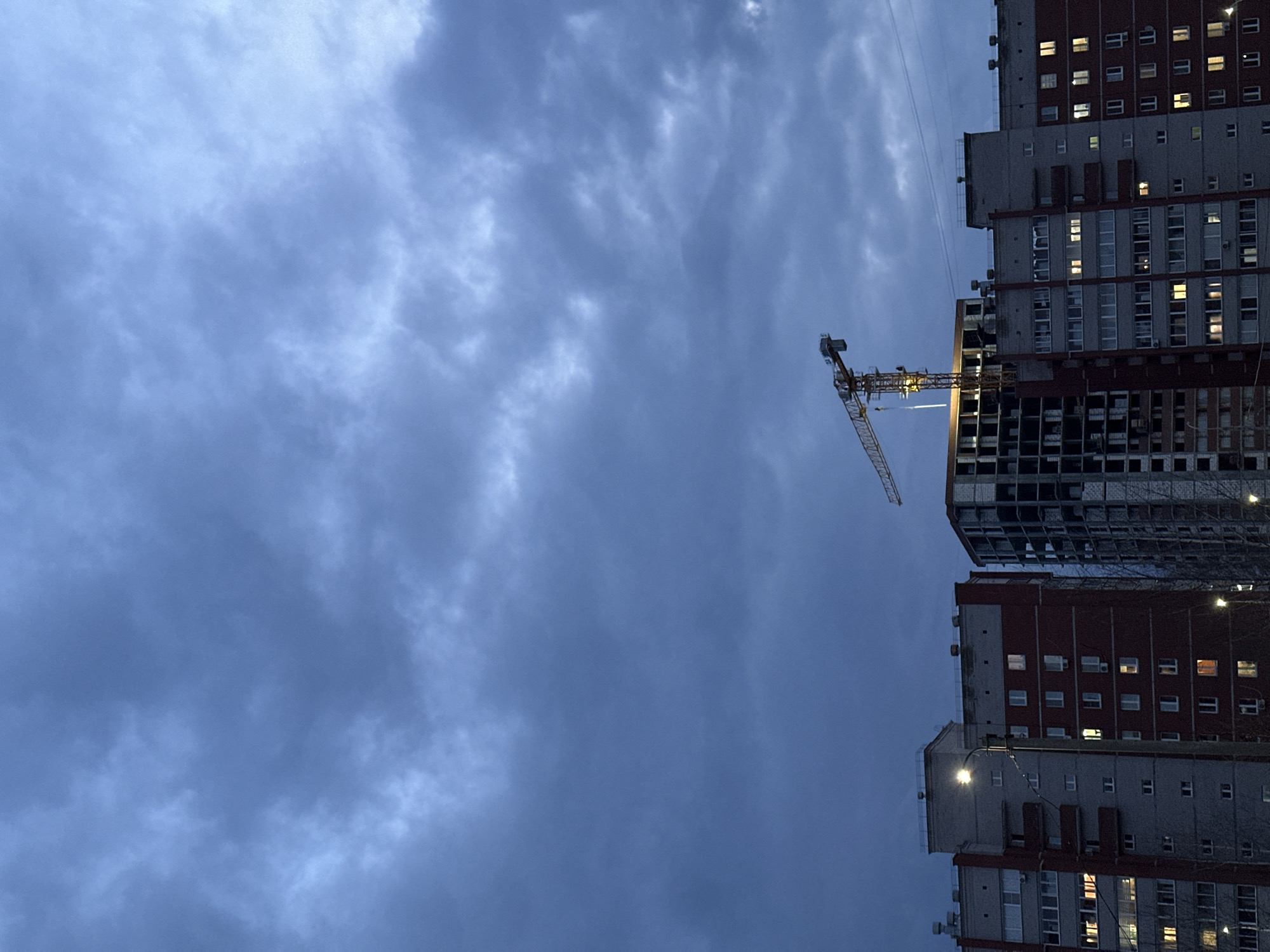 The image shows a construction site in Chaikovsky, Russia at dusk or dawn. A tall construction crane is positioned between two high-rise apartment buildings. One building is partially completed with exposed structural elements, while the other appears finished with lit windows. The sky is overcast with dark, textured clouds.
