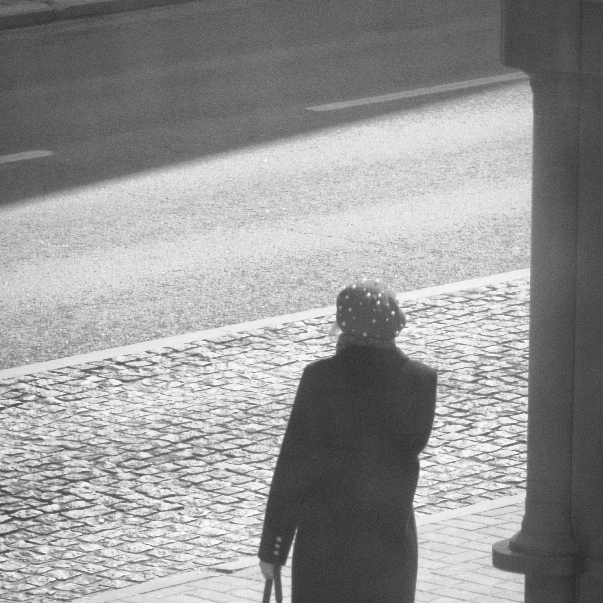 The monochromatic image, captured in the city of Hrodna, Belarus, depicts an outdoor scene featuring a single individual viewed from behind. The person is wearing a dark coat and a distinct light-colored head covering adorned with multiple circular embellishments. An indistinct dark object is held in the subject's left hand. The individual stands on a light-toned paved surface, which features an interlocking geometric pattern, positioned adjacent to a dark, vertical structural column or pillar that occupies the far right of the frame. A light-colored curb separates this paved area from a wider, darker asphalt road. A faint linear marking is present on the road surface. The scene is illuminated by strong directional light, evidenced by a prominent diagonal shadow extending across the road from the upper left.