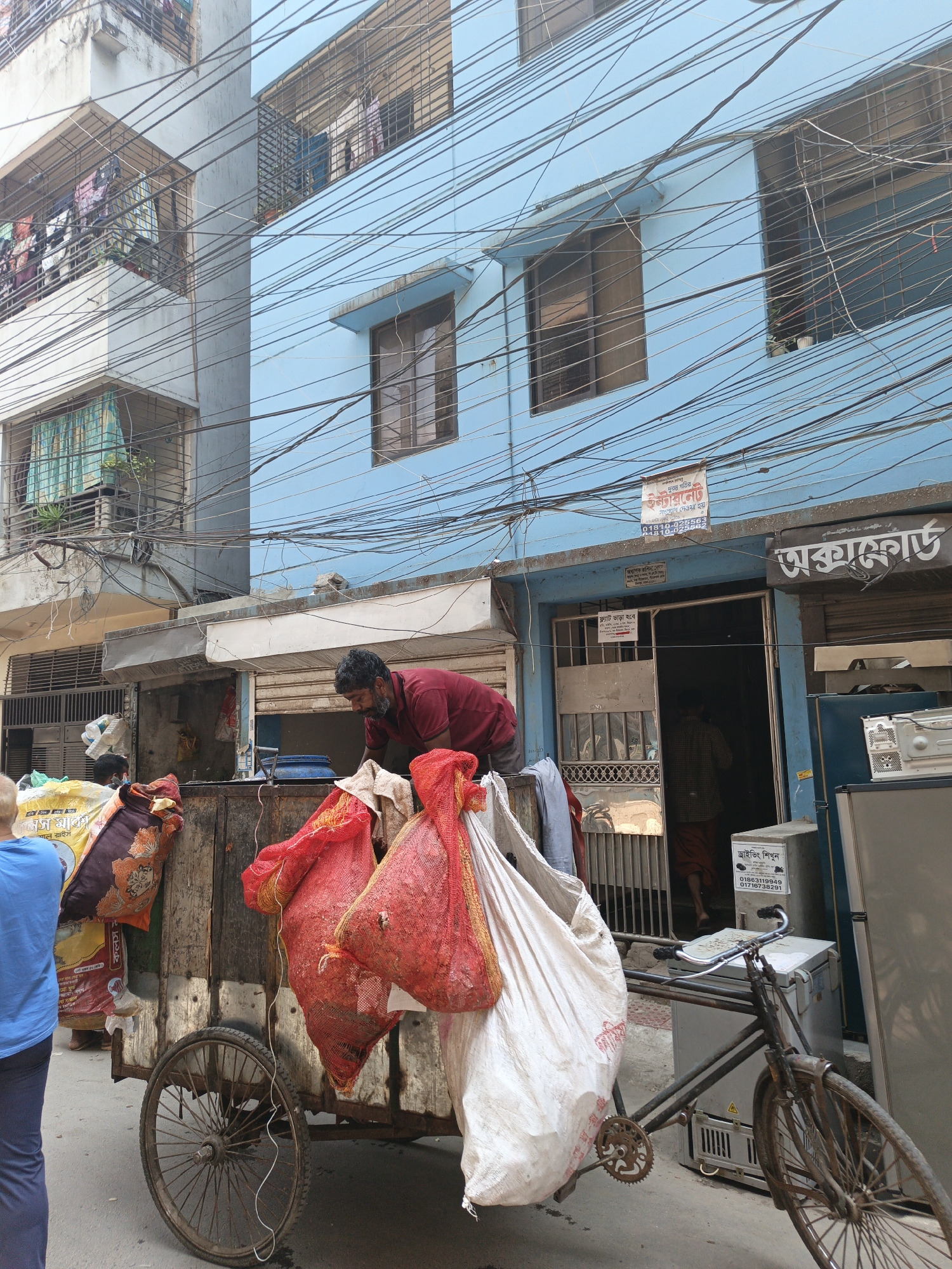 The image depicts a street scene in Dhaka, Bangladesh, with a man standing on a wooden cart filled with large bags of assorted items, including what appears to be refuse. He is wearing a red shirt and has a beard. The cart is attached to a rusty bicycle. The backdrop features buildings of different colors (white and light blue) with windows and power lines crisscrossing above. A shopfront with a sign in Bengali, identified with OCR as "অক্সাফোর্ড" (Oxford), is partially visible, along with advertisements, including one that reads "ফ্ল্যাট ভাড়া হবে" (Flat for rent) and "ড্রাইভিং শিখুন" (Learn driving).