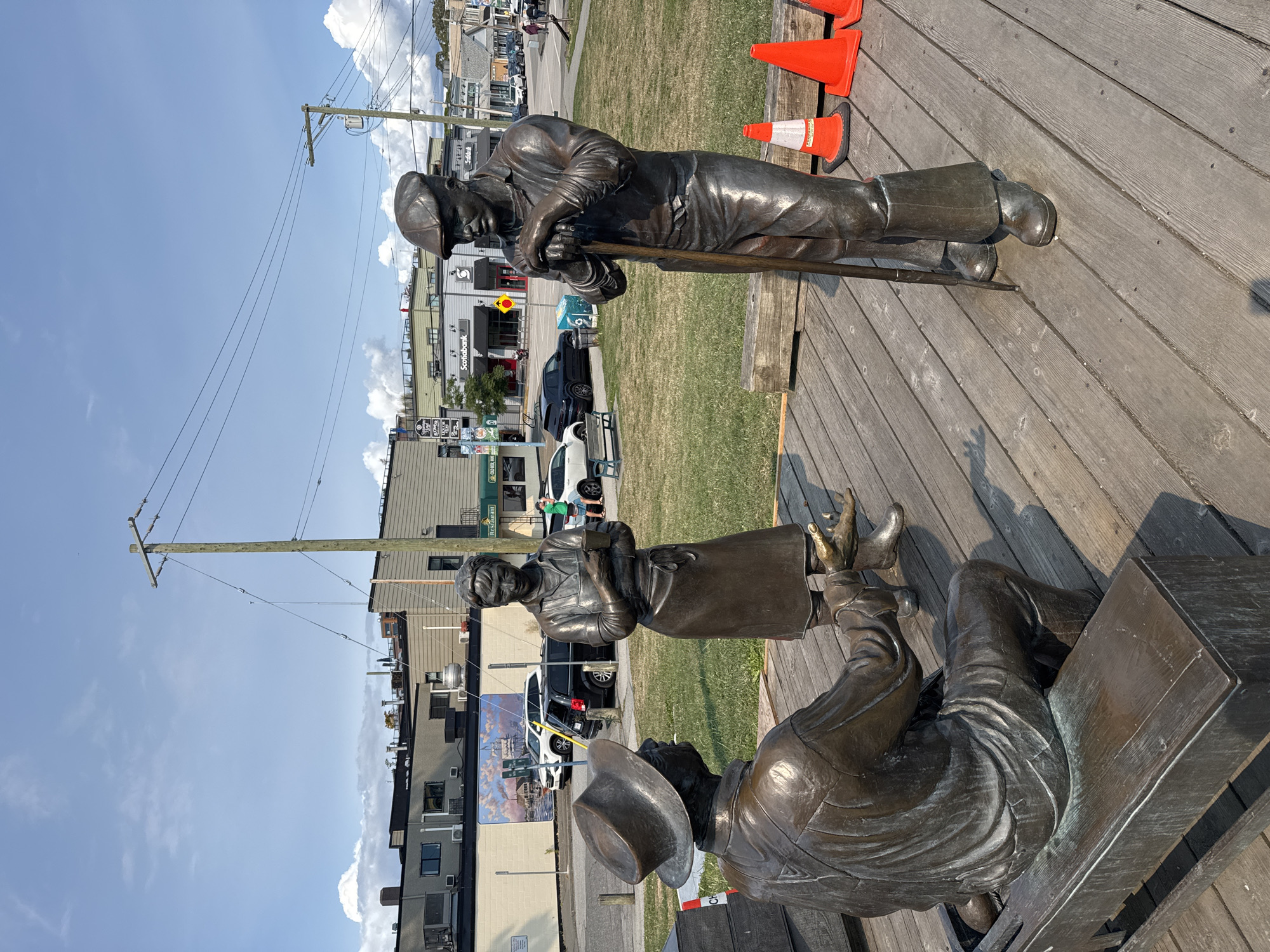 This is a photograph of a bronze statue depicting three people in a conversational pose, located in the city of Richmond, Canada. The statue is situated on a wooden deck. In the foreground, a man is seated on a bench, wearing a hat and overalls, and appears to be speaking. To his right, a woman in an apron and dress stands holding a mug, looking towards the seated man. To the right of the woman, a man wearing a flat cap and overalls stands leaning on a shovel, facing the other two figures. Behind the statue, there is a grassy area with some orange traffic cones and a wooden barrier. Further back, a street with buildings, cars, and pedestrians can be seen. The buildings are of varying heights and architectural styles, with some featuring signage such as "Scotiabank." The sky above is partly cloudy.