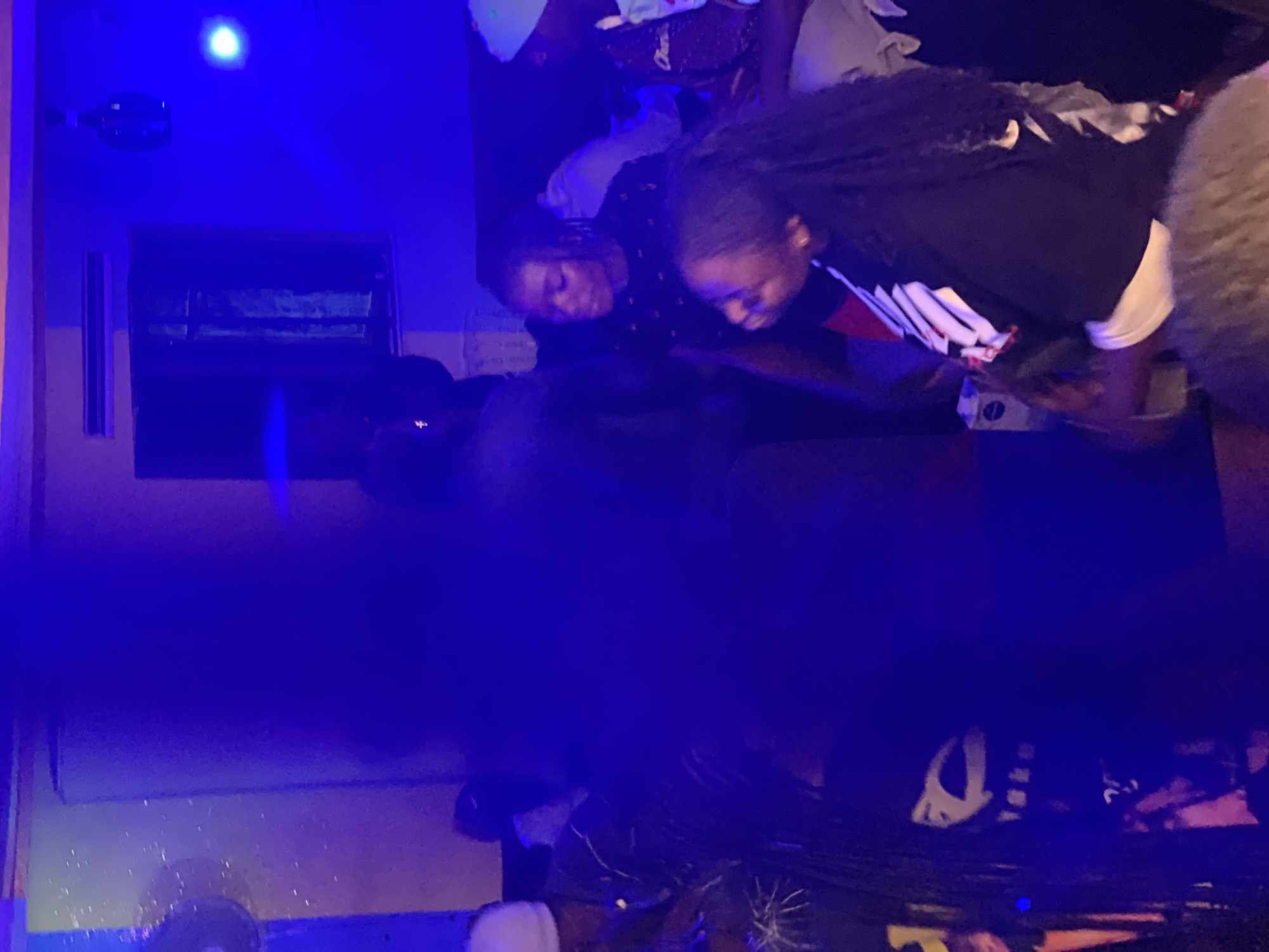 The image depicts a dimly lit indoor scene, possibly a club or social gathering, bathed in vibrant blue and purple lighting. The setting appears to be Durumi, Nigeria. In the foreground, several individuals are visible, their features somewhat obscured by the low light and motion blur. To the left, a person wearing a white head covering is partially visible. Central to the image, a woman with dark hair pulled back is seen looking downwards, dressed in a dark, patterned top. Beside her, another individual, also with dark hair, is wearing a black shirt with white and red graphic elements. In the background, more people are present, some appearing to be dancing or socializing. The environment features a dark ceiling with what appears to be a window or ventilation unit, and a bottle suspended from the ceiling, illuminated by a blue light. The overall atmosphere is lively and dynamic, indicative of a social event.