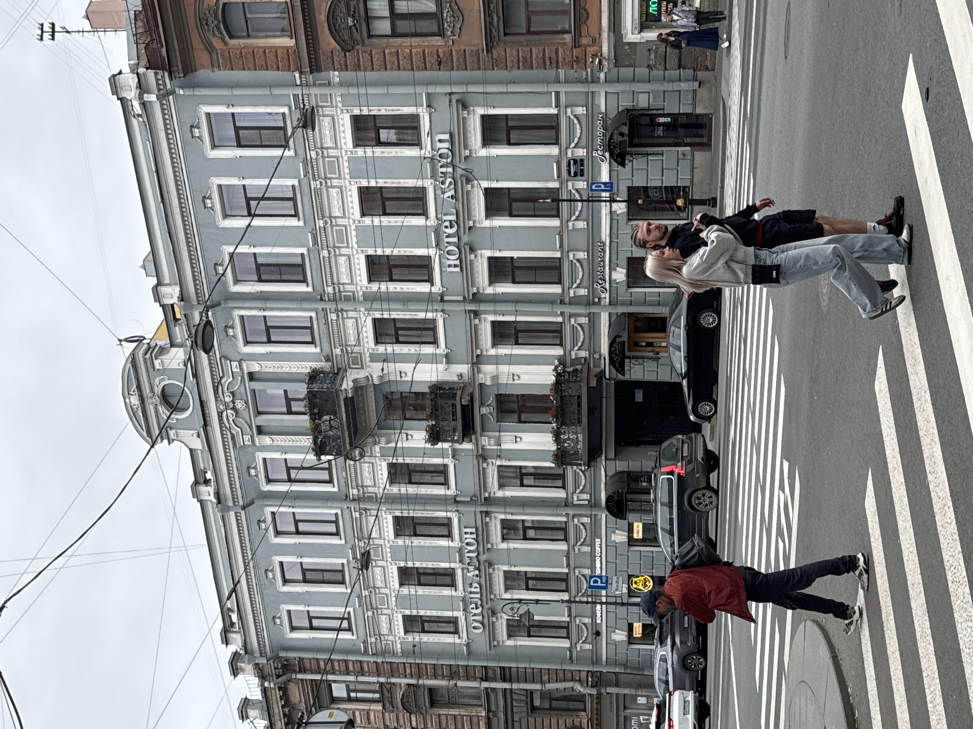 The photograph shows a street scene in Saint Petersburg, Russia. "Hotel Aston" is prominently displayed on the facade of a multi-story building with ornate window frames. A couple is crossing a pedestrian crosswalk. Another individual with a red jacket is also crossing. Cars are parked along the street. Overhead power lines run across the building.