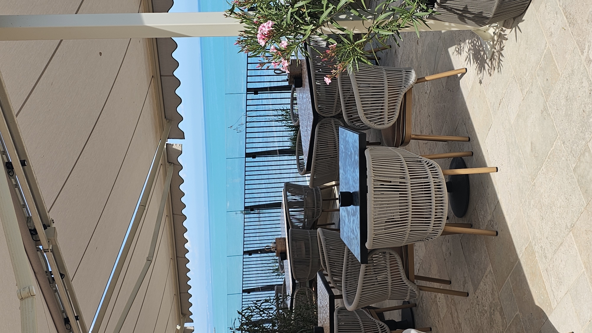 An outdoor cafe with a view of the sea is shown in Varna, Bulgaria. The cafe is furnished with tables and chairs, and there are plants and flowers. The sea is a bright blue, and the sky is clear. The overall atmosphere is relaxed and inviting.