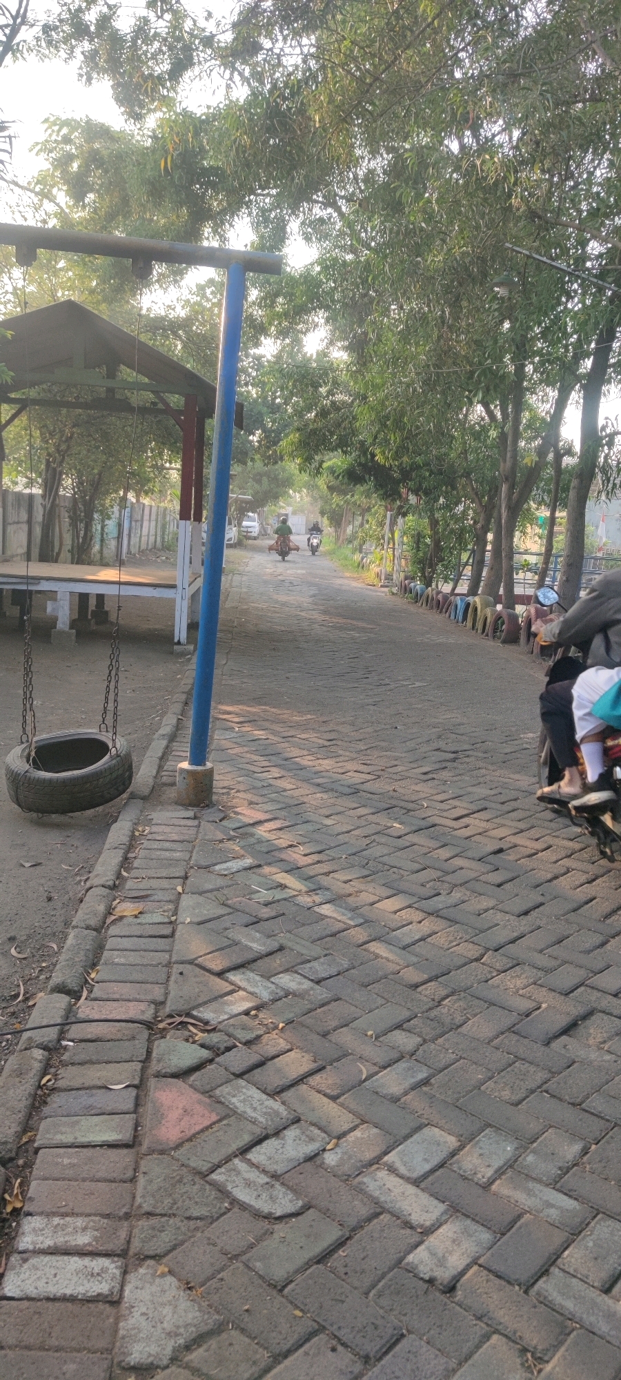 The image depicts a paved road in Tebel, Indonesia, likely a park or recreational area given the presence of a tire swing and a covered seating area. The road is constructed with interlocking brick pavers laid in a herringbone pattern. Tall trees with dense foliage line the right side of the road, casting shadows on the pavement. On the left side, there is a covered structure with a raised wooden platform and a tire swing suspended by chains from a blue pole. A fence made of concrete blocks runs along the left edge of the property, with vegetation behind it.
Several individuals on motorcycles are visible on the road. One motorcycle in the foreground is partially visible from the rear, with two people seated on it. Further down the road, two more motorcycles are heading away from the viewer. The atmosphere suggests a late afternoon or early morning setting due to the low angle of the sunlight and the long shadows. The overall scene is one of a quiet, outdoor public space.