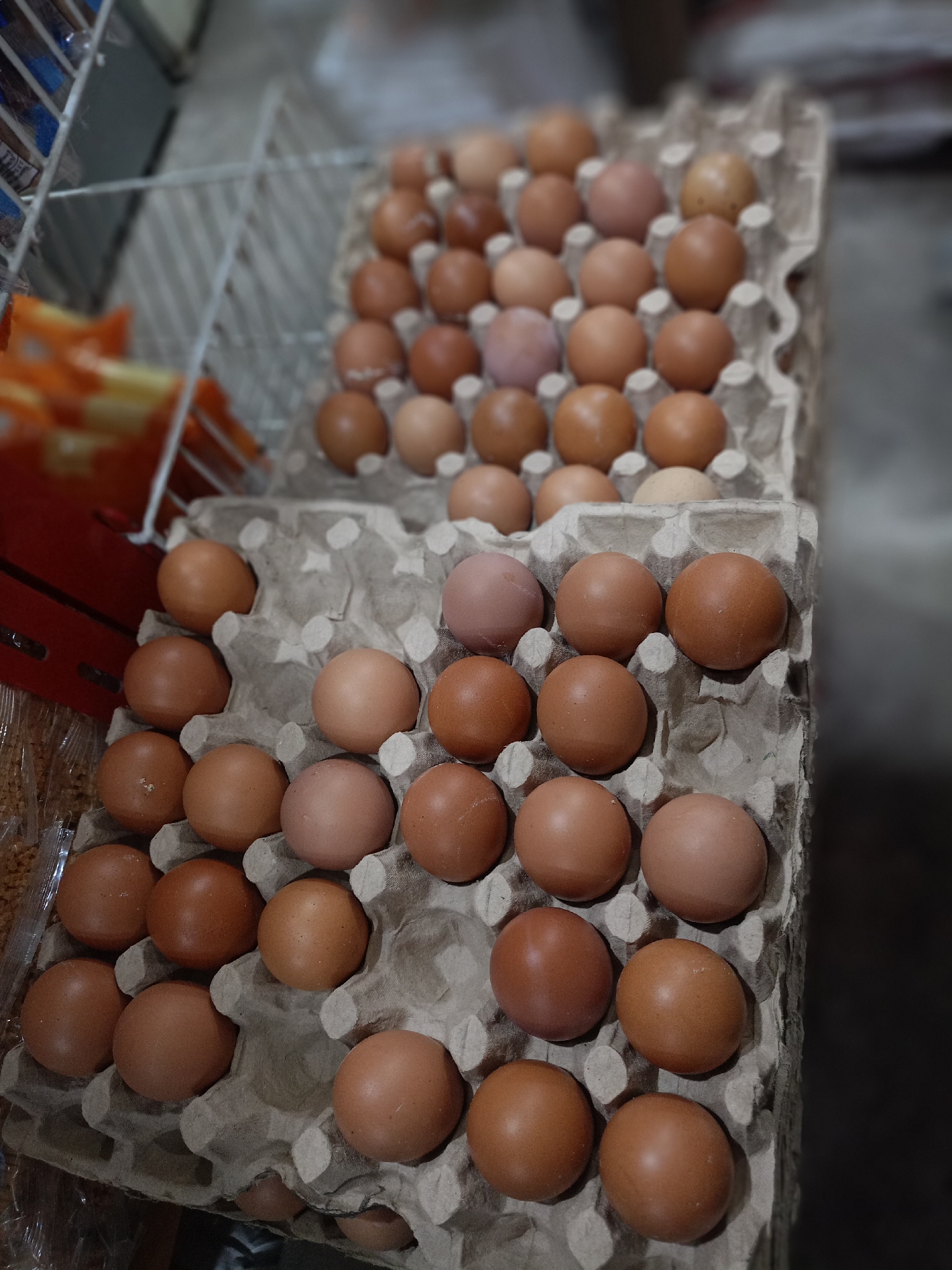The image depicts two stacked egg cartons filled with brown eggs. The location is Uteunreutoh, Indonesia. The cartons are made of molded pulp. The eggs vary slightly in shade. A wire basket and red plastic container are visible on the left edge. There are no people visible in the frame.