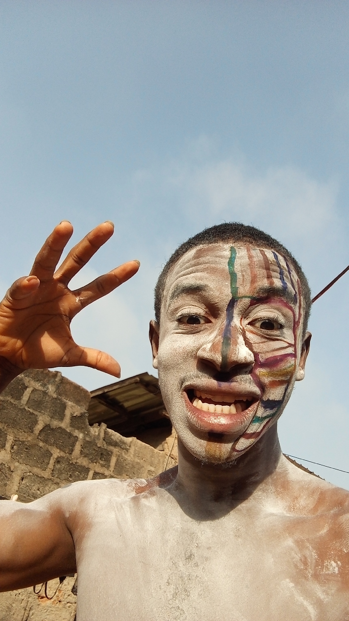 A shirtless man, his skin covered in white powder and face painted with colorful lines, is posing outside under a blue sky in Onitsha, Nigeria. His teeth are visible in a wide smile. His right hand is raised, with fingers spread apart. The background includes a brick wall and a corrugated metal roof. A wire is visible in the top right corner of the frame.