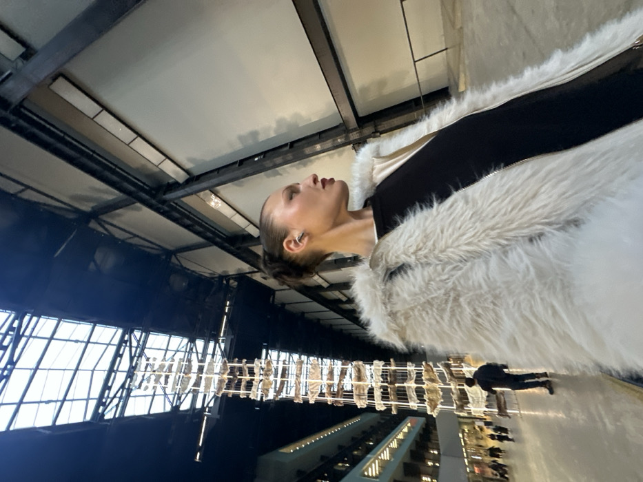 From a low angle, a woman in a furry white coat with black sleeves looks upward. Architectural details include steel beams and gridded windows. Below, a walkway with people and modern architecture are visible. A hanging sculpture casts reflections on the floor. The woman wears an ear cuff and has her hair pulled back.