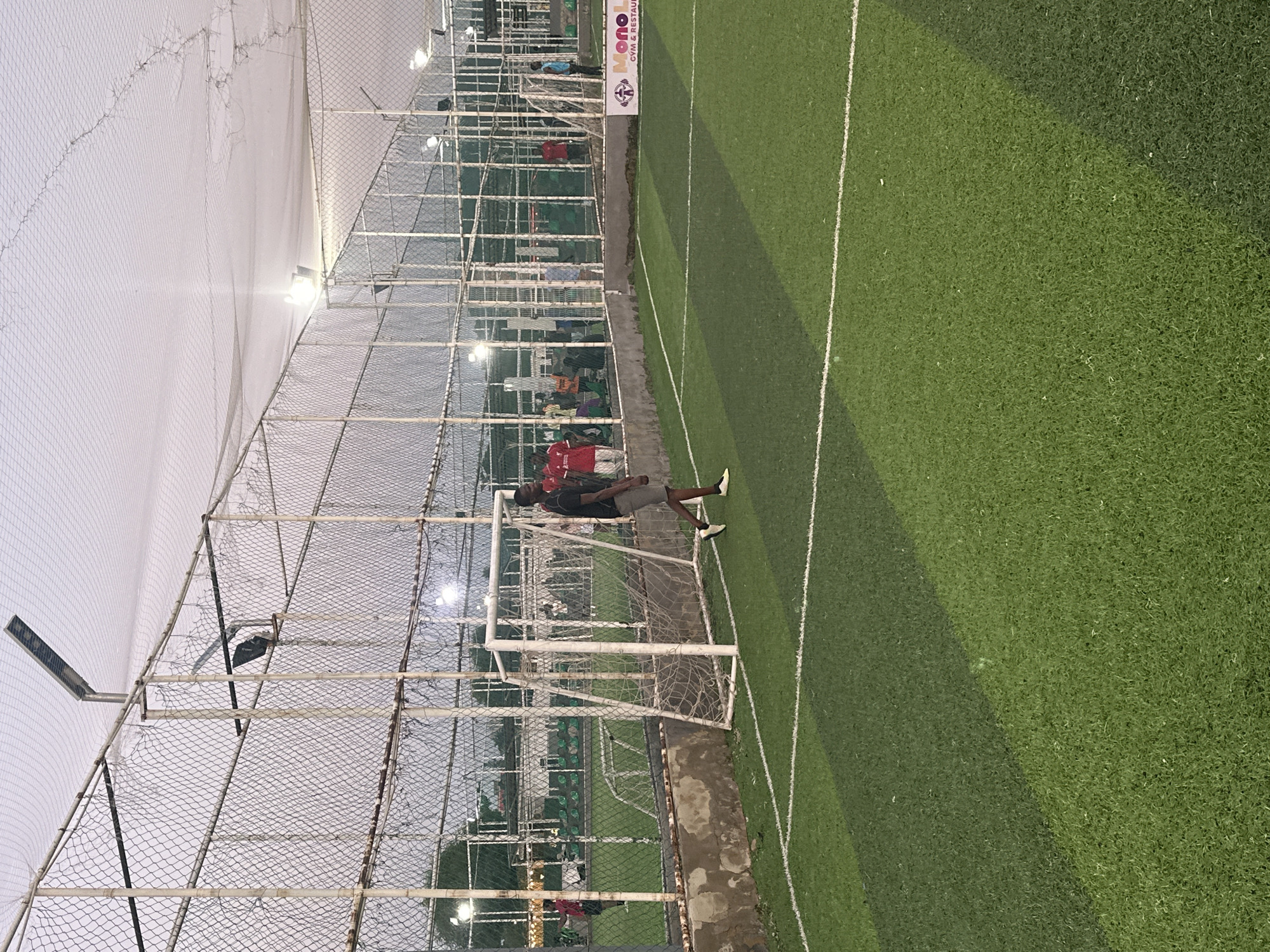 This is an outdoor photograph of a futsal pitch located at Mono Leisure in Abuja, Nigeria. The pitch has a green artificial turf surface with white markings. A white futsal goal is visible on the right side of the frame, with a net. In the foreground, a Black male wearing a dark t-shirt and grey shorts walks across the pitch. In the background, several other people are present, some wearing red shirts. The pitch is enclosed by a fence with a mesh netting on top, and artificial lighting fixtures are visible. The sky is overcast. A banner for "Mono Leisure" is attached to the fence on the right side.