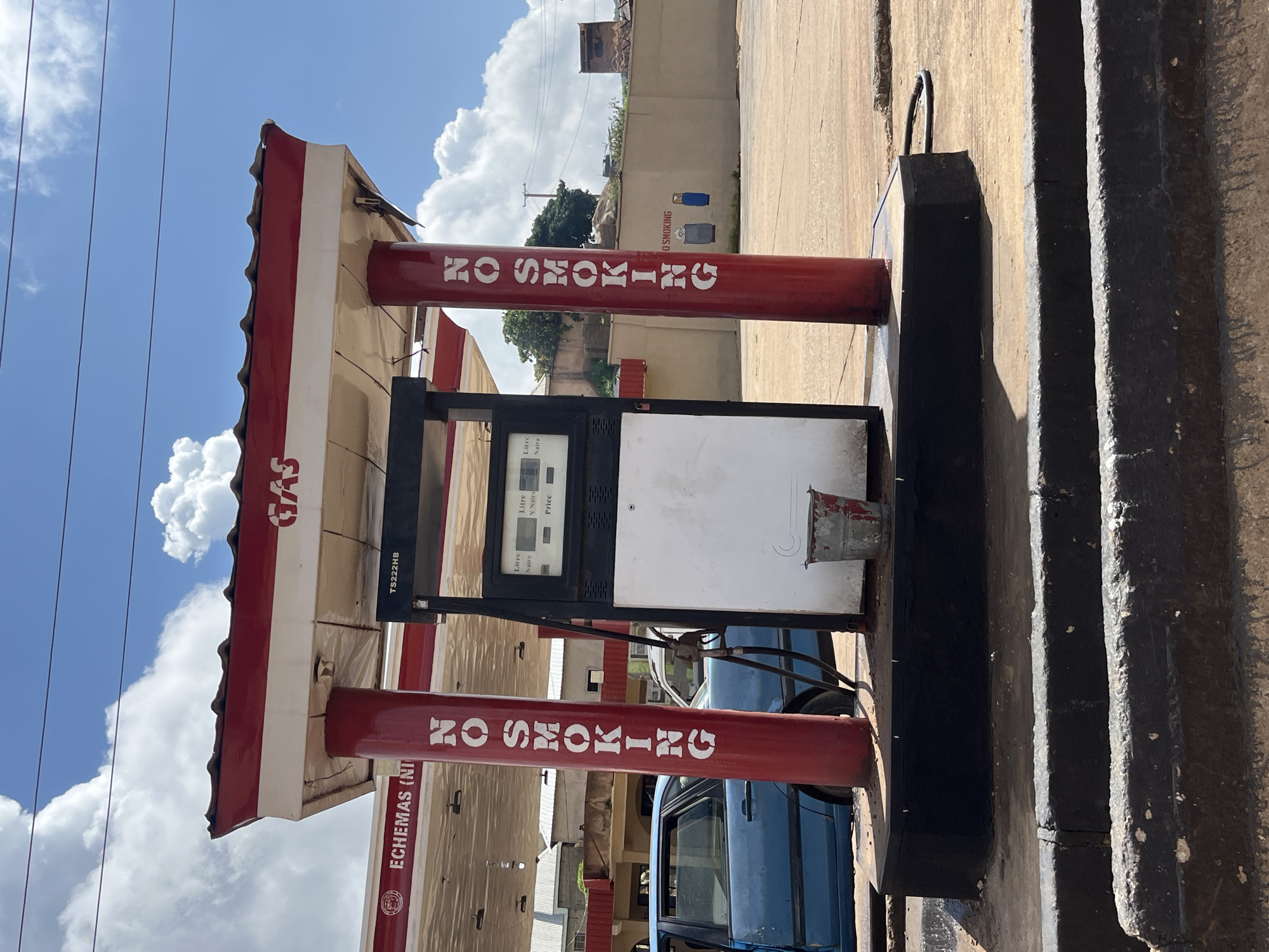 The image depicts a gas station in Jos, Nigeria, under a partly cloudy sky. The central focus is a red and white gas pump station with a red tiled roof. The pump itself is black and white, displaying readings for "Litre," "Naira," and "Price." On the red support columns of the station, "NO SMOKING" is prominently displayed in white lettering. To the left, the name "ECHMAS (NT)" is visible on an awning.
A light blue sedan with its doors open is parked next to the gas pump. In the background, a light brown wall with a mural of a gas cylinder and a blue object is visible, along with distant buildings and trees. A small, red-rimmed metal bucket sits on the ground near the base of the gas pump. The ground surface appears to be paved concrete, with a black curb in the foreground. Electrical wires are strung across the top of the image. The overall scene suggests an operational but somewhat aged gas station.