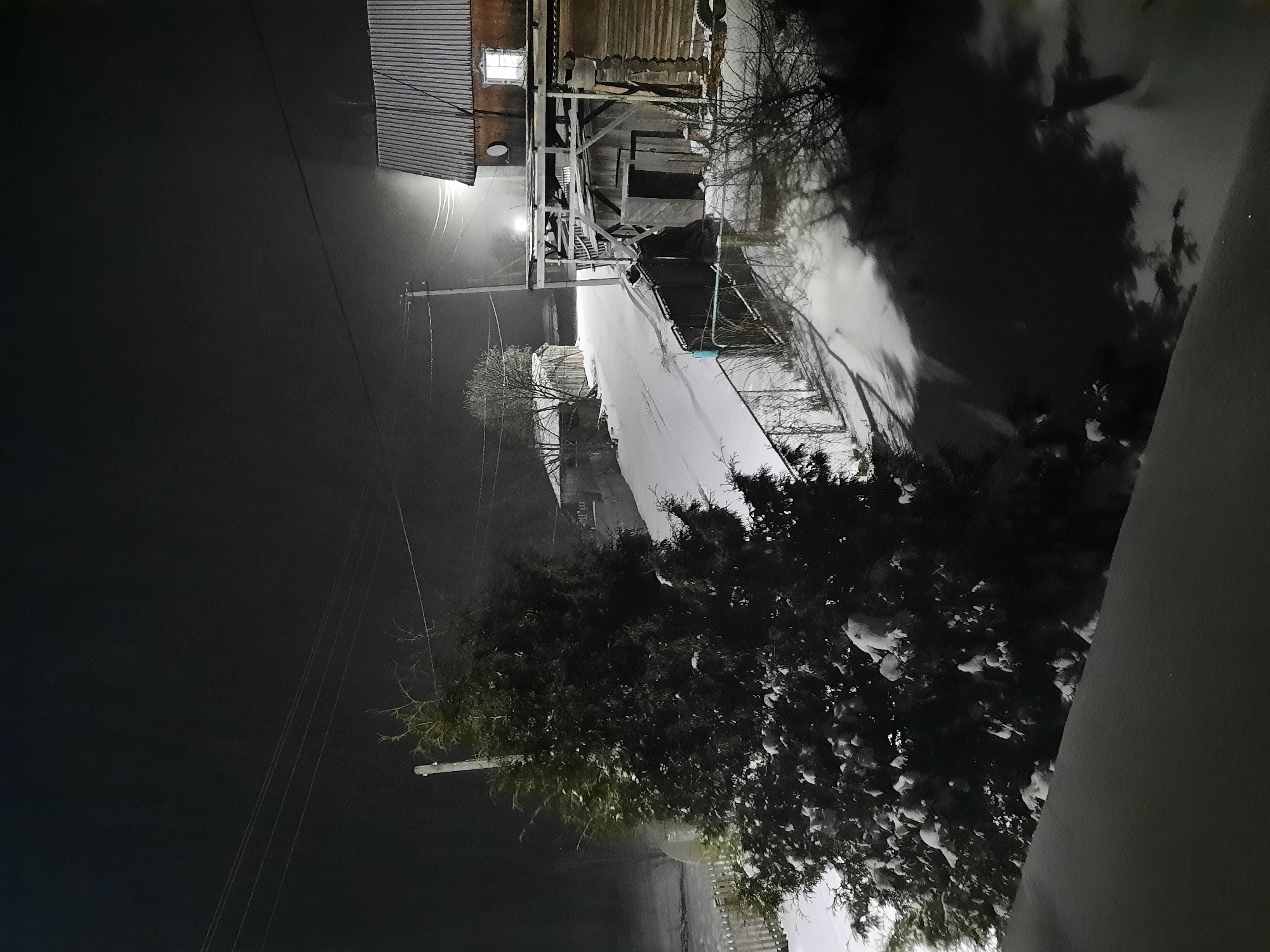 The image depicts a snowy street scene at night in what appears to be a residential area in Архангельское, Russia. A street light illuminates the snow-covered road, houses, and trees. The houses are simple, likely older constructions with visible eaves and possibly metal roofs. Power lines are visible crossing the dark sky on the left side of the image. A large, dark bush with snow on it is in the foreground. The scene is quiet and still, suggesting a cold winter night. There are no people visible in the image.
