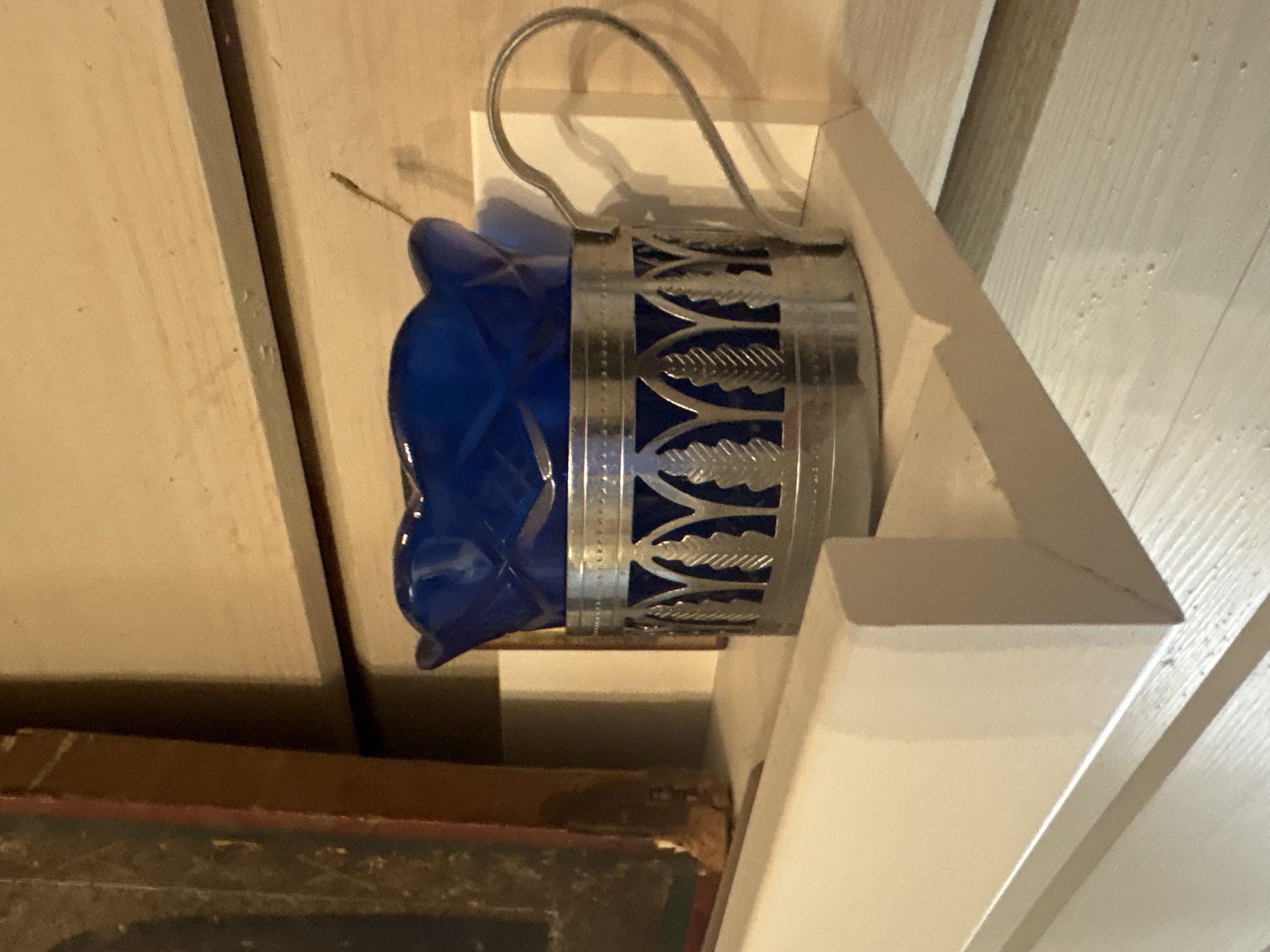 A cobalt blue glass, likely a candle holder or a small vase, is placed in a silver metal holder with an ornate design. The holder has a handle and decorative cutouts resembling leaves. The glass itself features a diamond-patterned texture. This ensemble sits on a light-colored shelf, which is part of a wooden wall. To the left of the glass holder, a portion of a dark-framed object, possibly a painting or a book, is visible. The background appears to be a light-colored wooden surface. The scene is lit, casting shadows that highlight the textures and forms of the objects. The location is identified as Березка Снт (поварово), Russia.